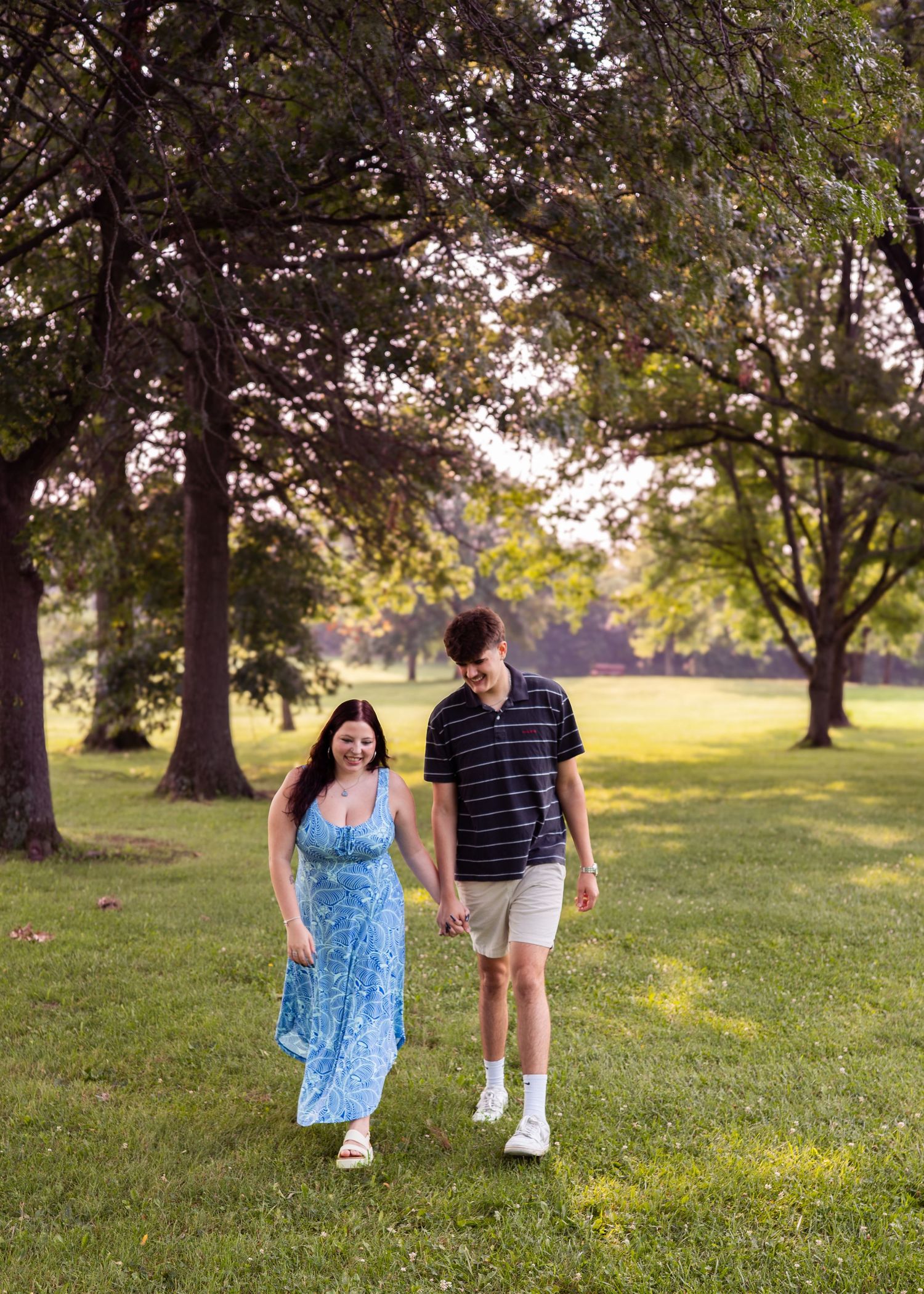 A couple walks hand-in-hand through a lush green park, the woman in a blue floral dress and the man in a striped polo shirt.