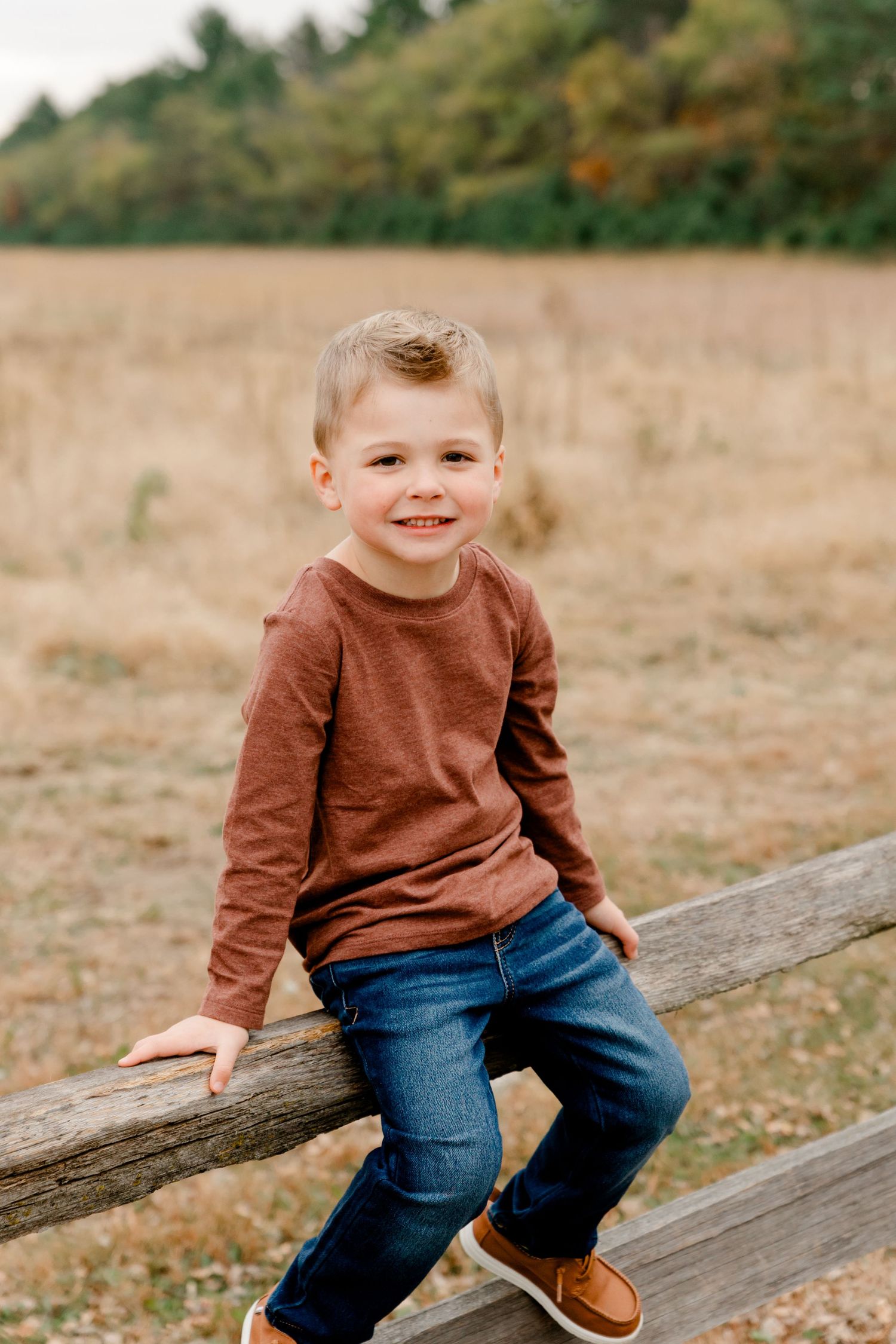 Happy young boy in rust long sleeve shirt sitting on wooden fence in fall field.