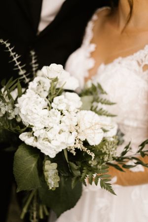 A wedding bouquet of white flowers and greenery held against a white dress.