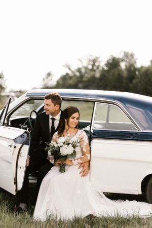 Wedding couple poses beside a classic white vintage car holding a beautiful white floral bouquet.