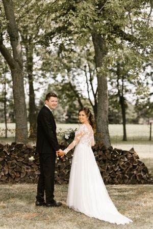 Full-length wedding portrait of a couple standing by a woodpile in a rustic outdoor setting.