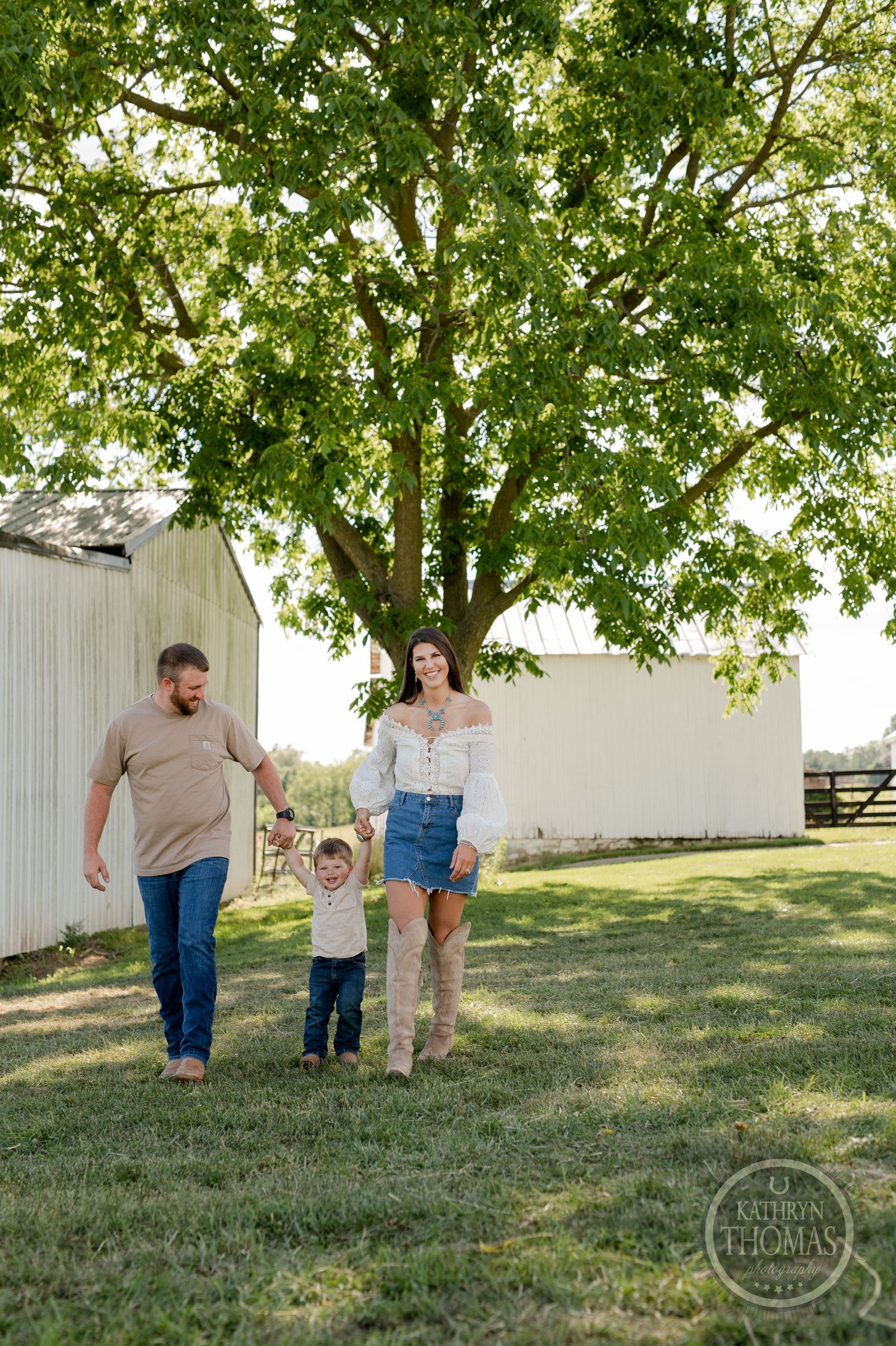Family enjoying walk on grassy field with white barn and leafy tree in background.