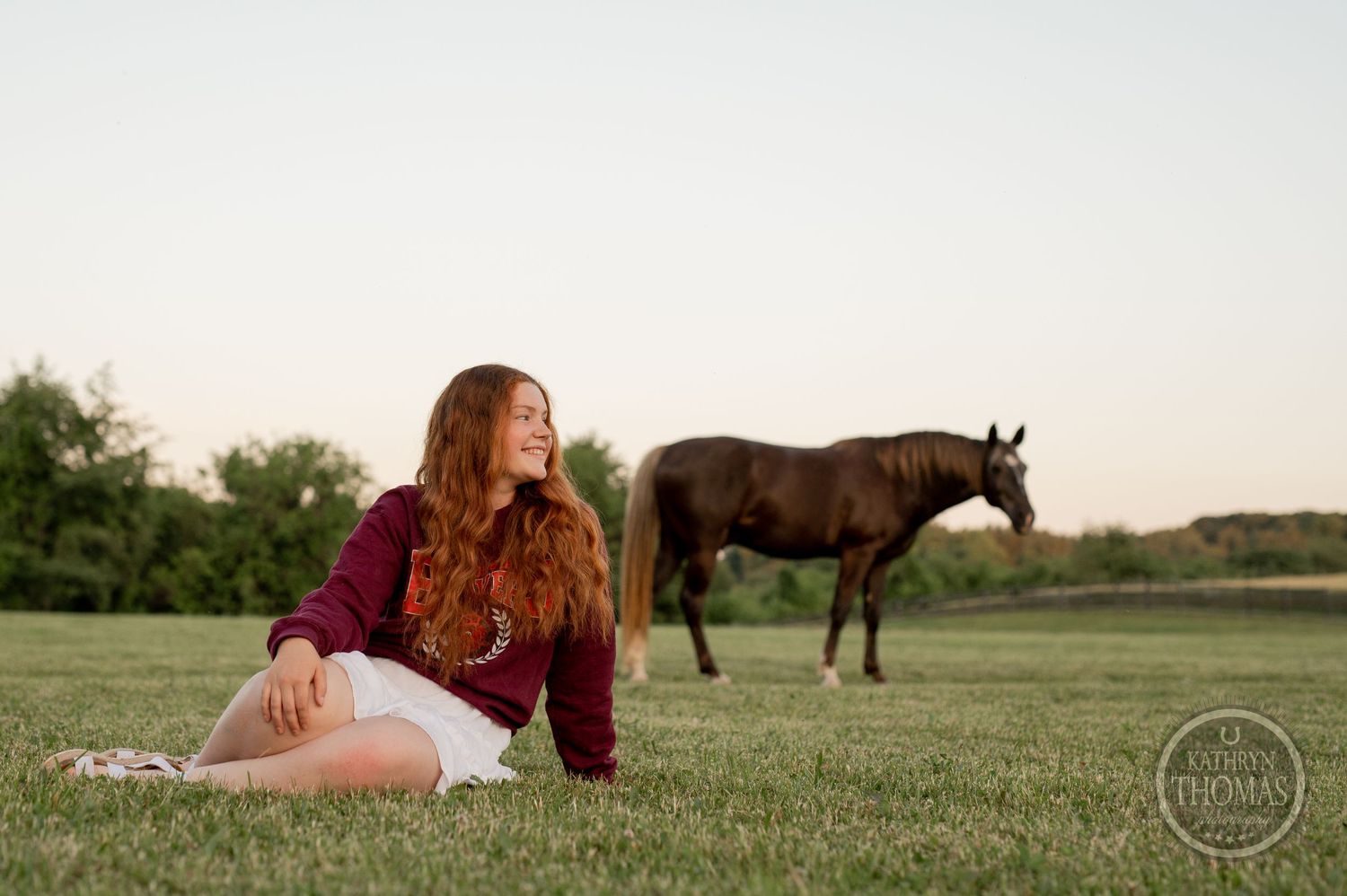 Person in burgundy sweater sits in grass with dark horse grazing in background.