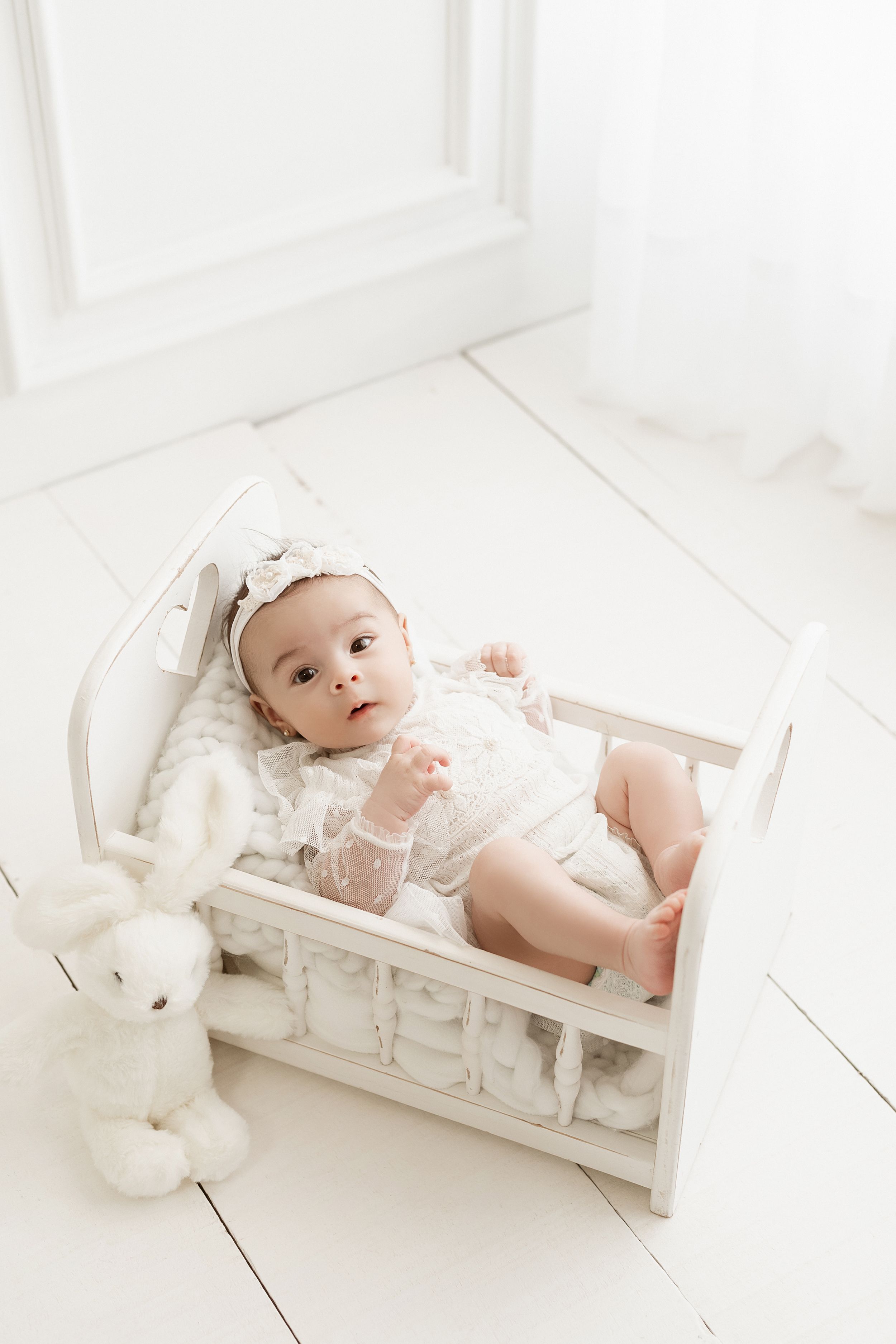 A small baby lies in a white wooden doll bed with a plush bunny toy against a white wall.