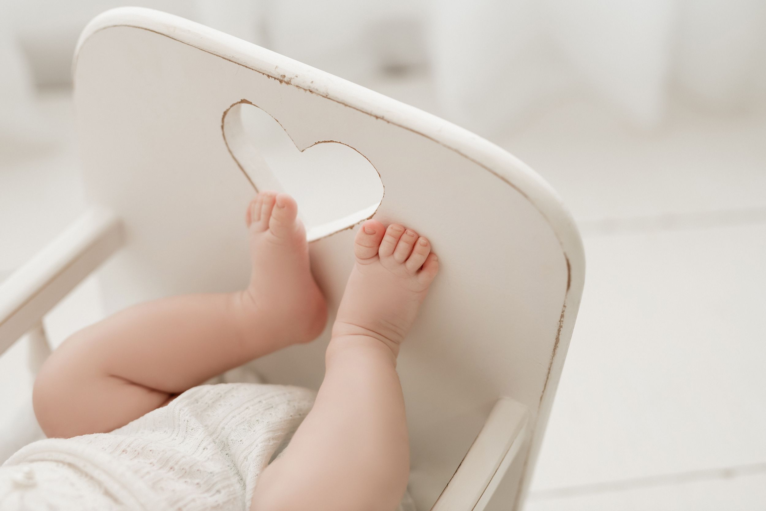 Tiny baby feet rest against a white chair with a heart-shaped cutout detail.