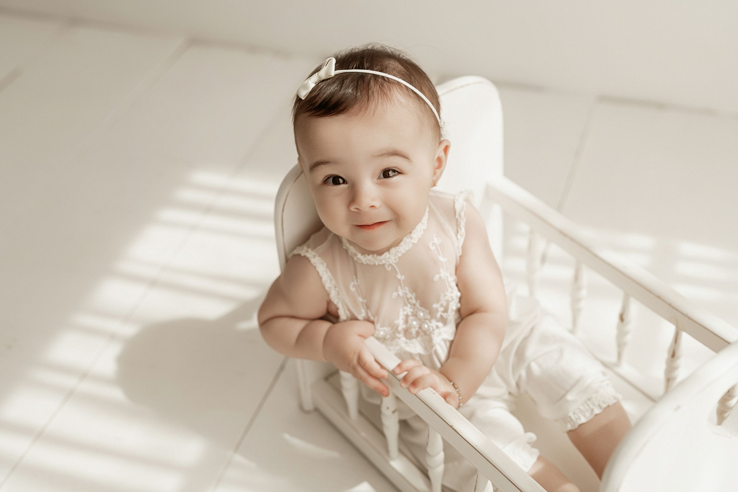 A small baby in a white lace dress sits in a white crib with sunlight streaming through window blinds.
