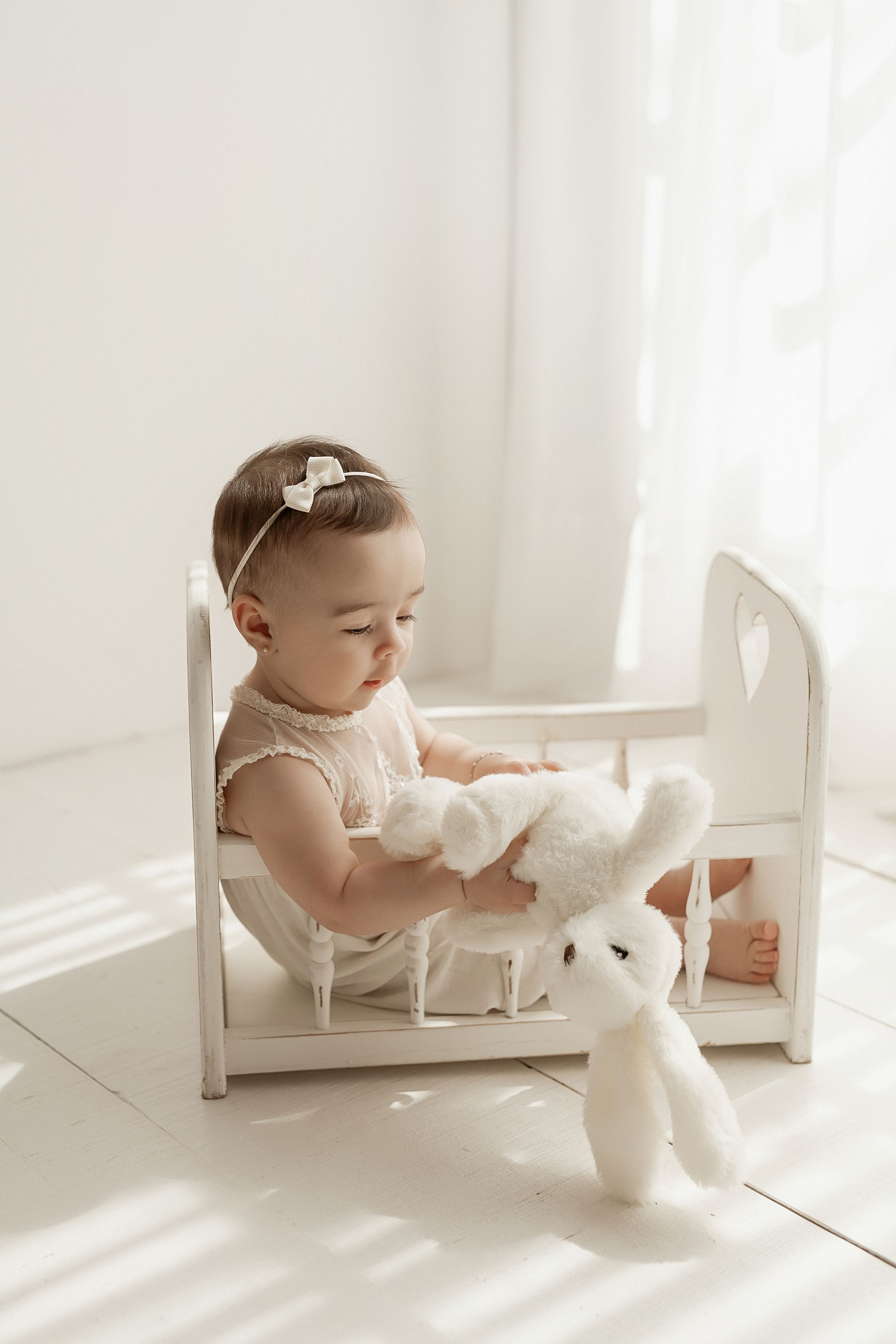 Toddler in white dress plays with plush bunny toy while sitting in small white wooden chair by window.