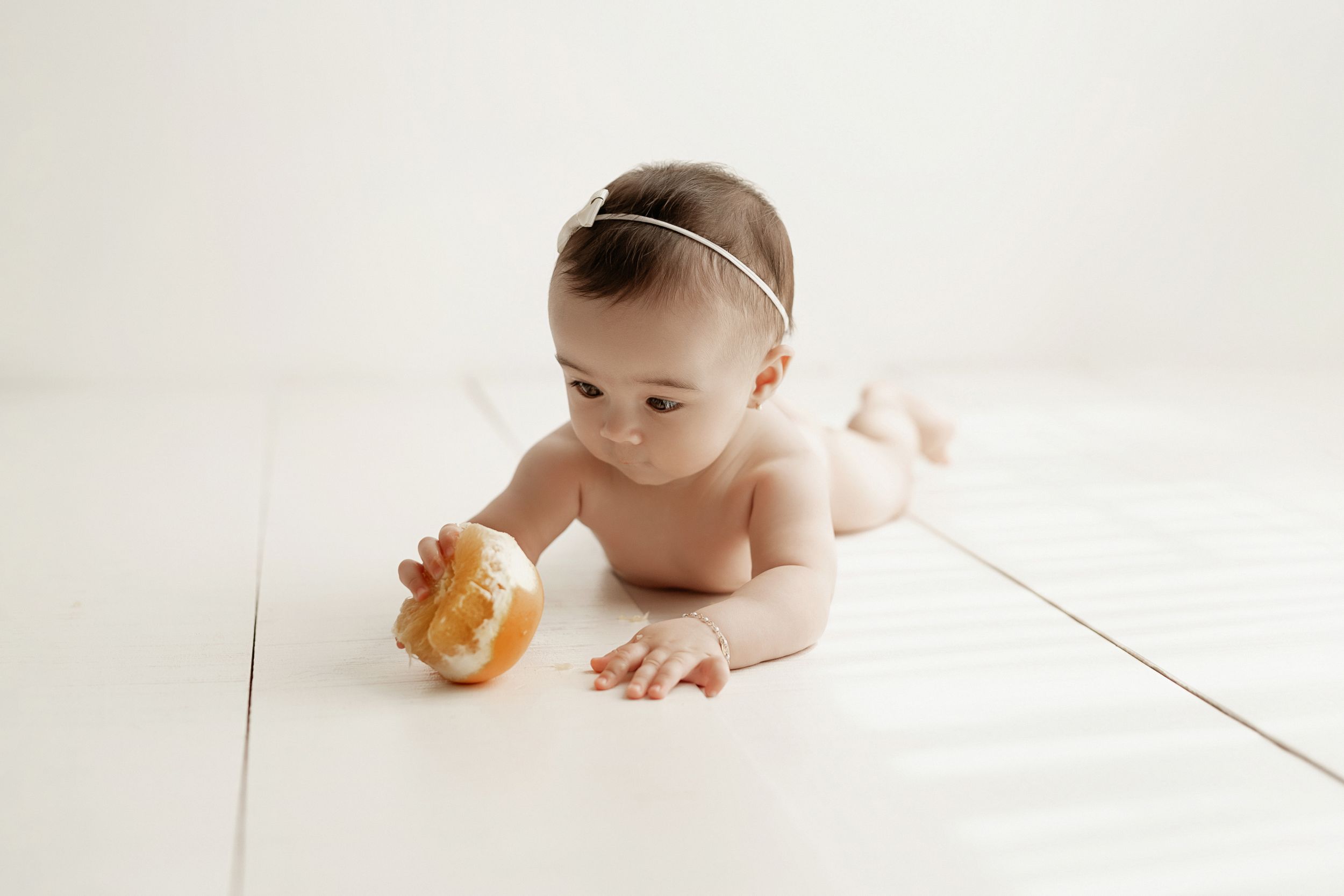Baby in white headband lying on stomach with orange toy on light background.