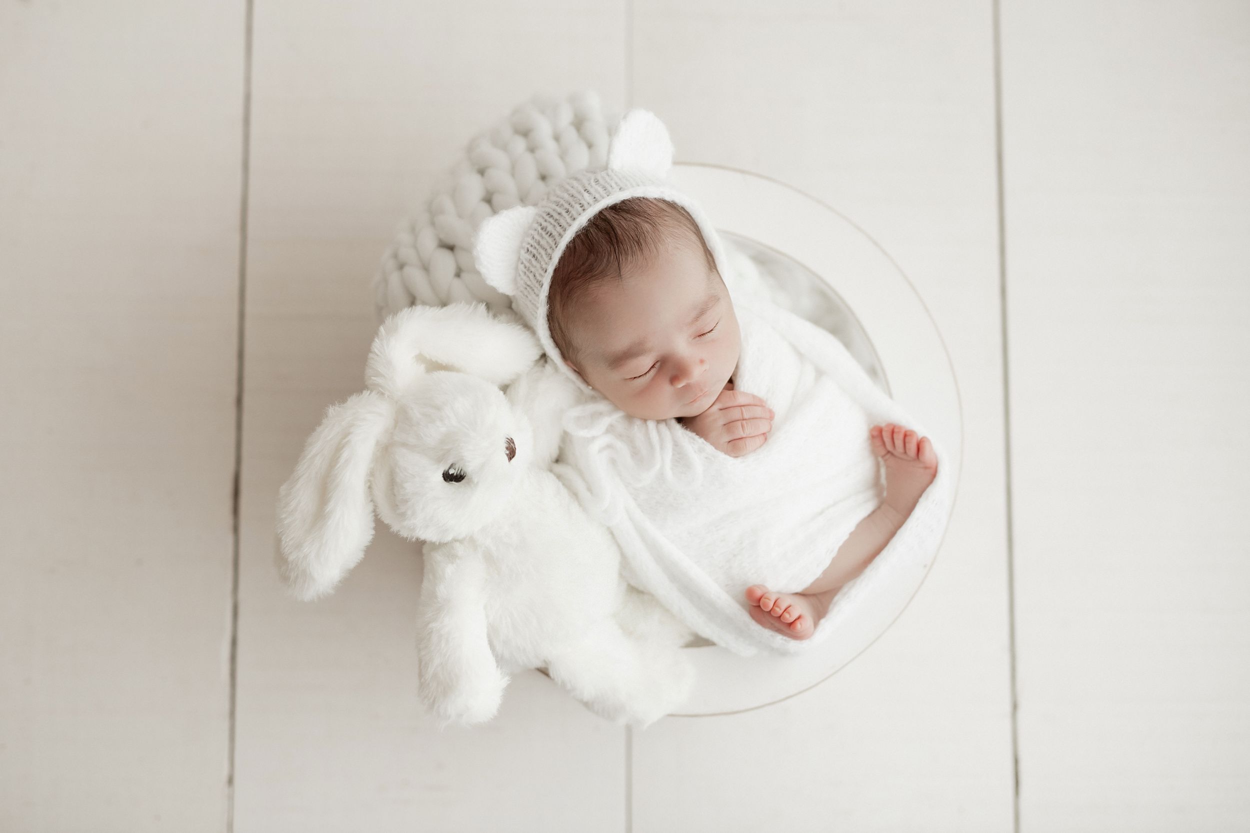A sleeping infant nestled in a white basket with a plush bunny toy on a light floor.