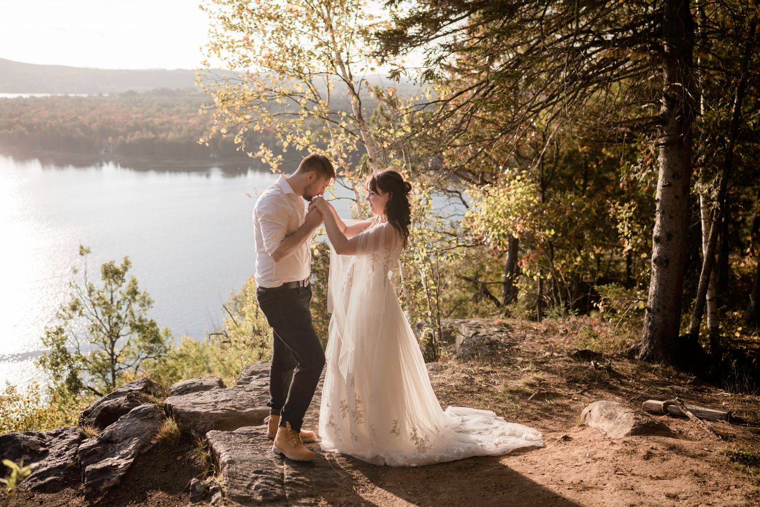 Eloping couple standing on a cliff, overlooking a lake. He kisses her hand in Mont Tremblant, Quebec, Canada.