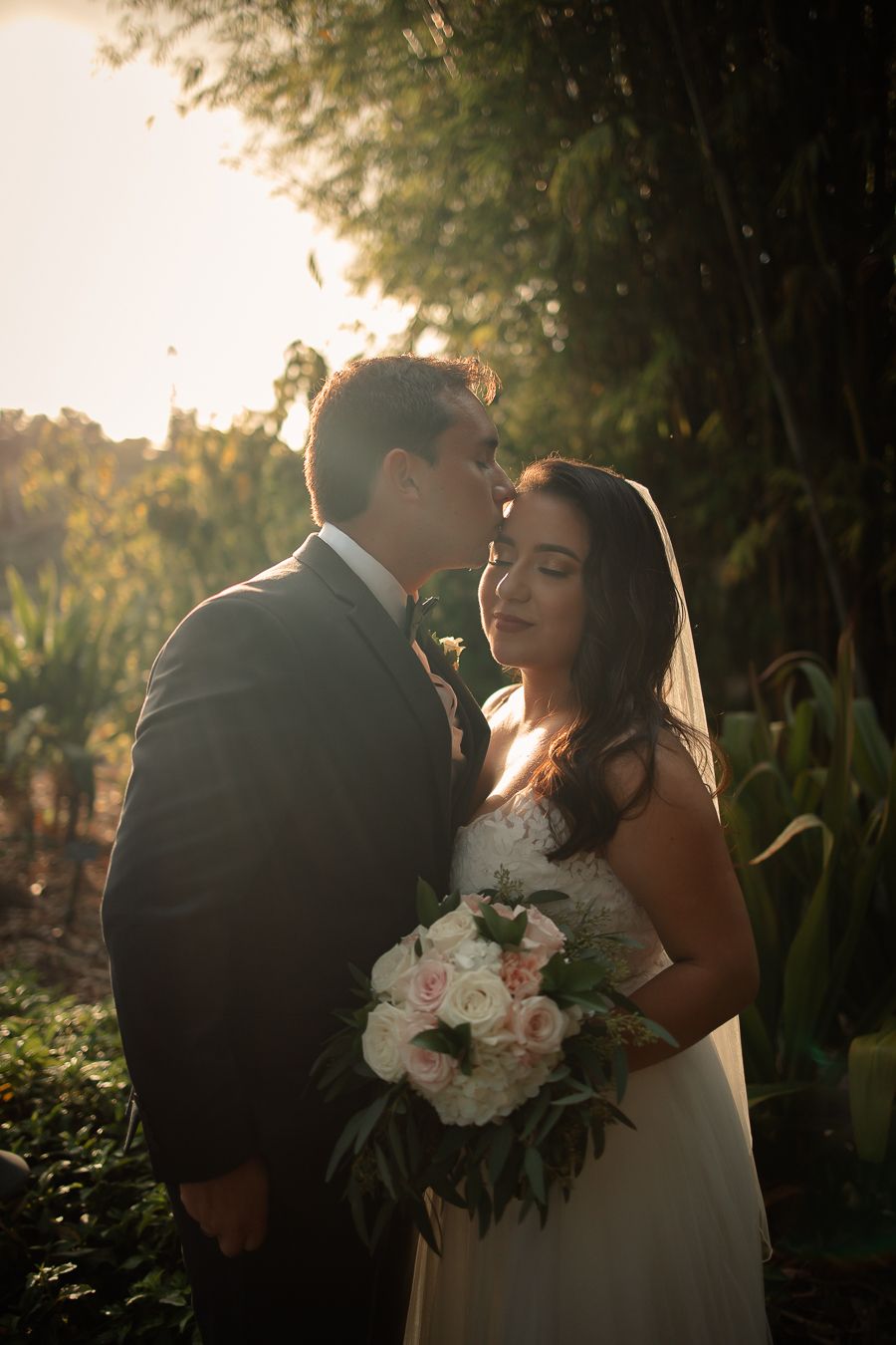 Groom kisses bride forehead in golden sunlight Romantic outdoor wedding photography capturing soft, intimate love with elegant bridal detail