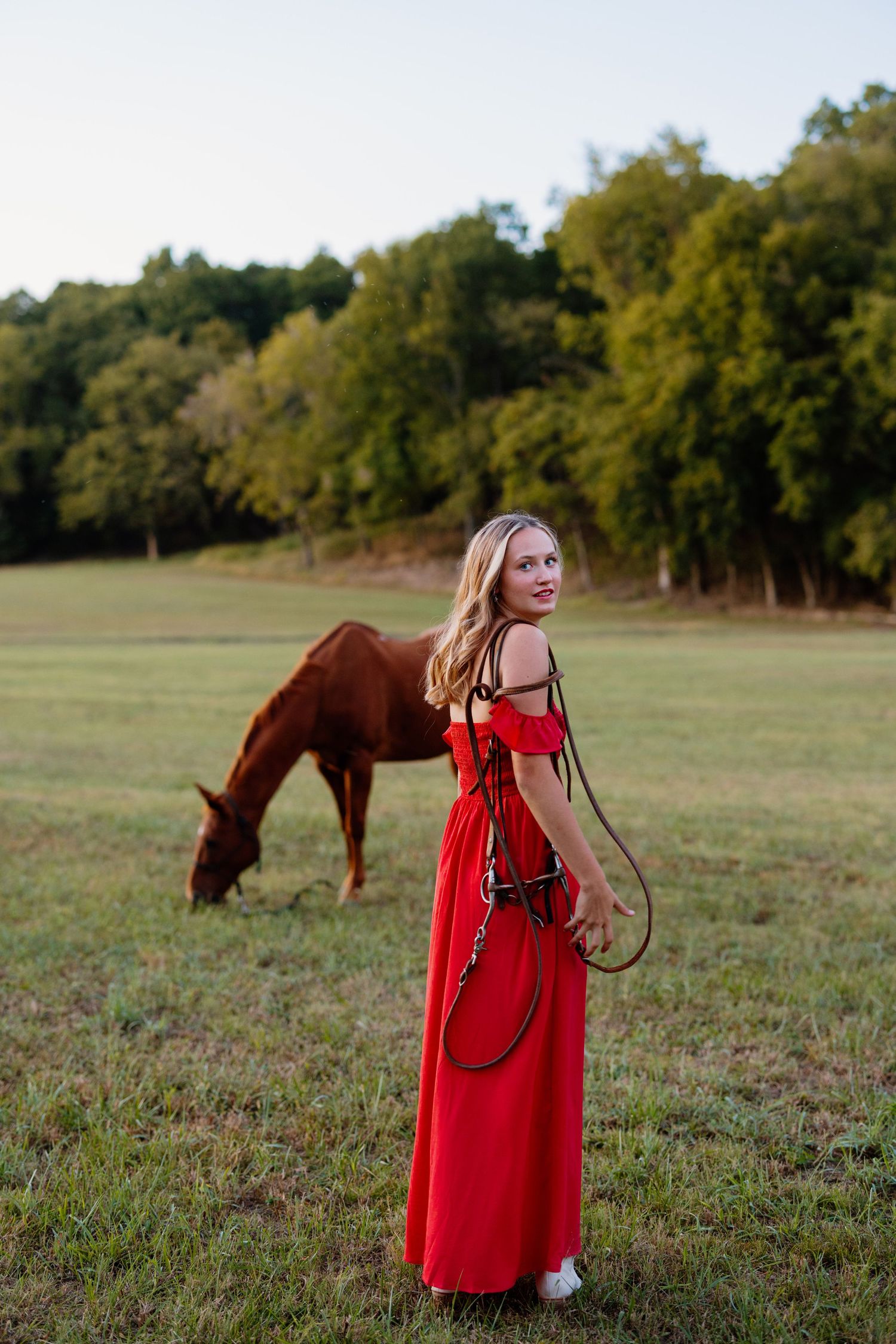 Woman in flowing red dress stands with grazing horse in scenic meadow at sunset.