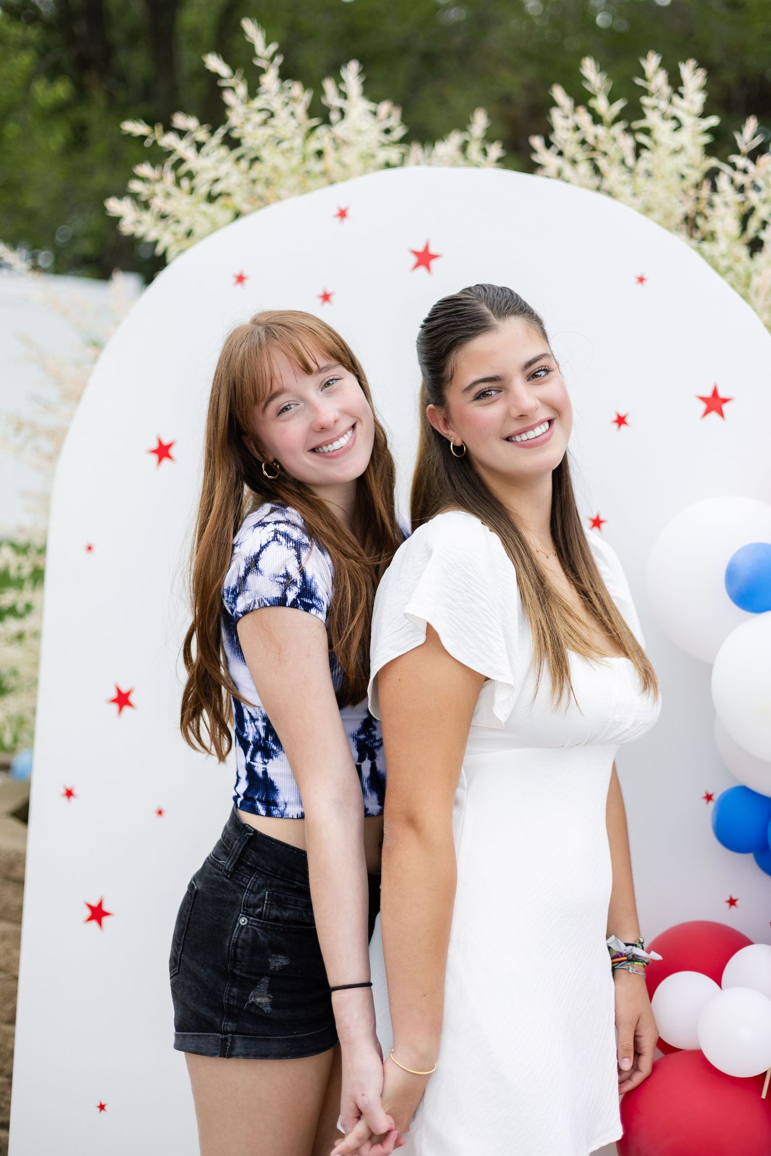 Two young women pose back-to-back against a white decorative backdrop with red stars and balloons at a celebration.