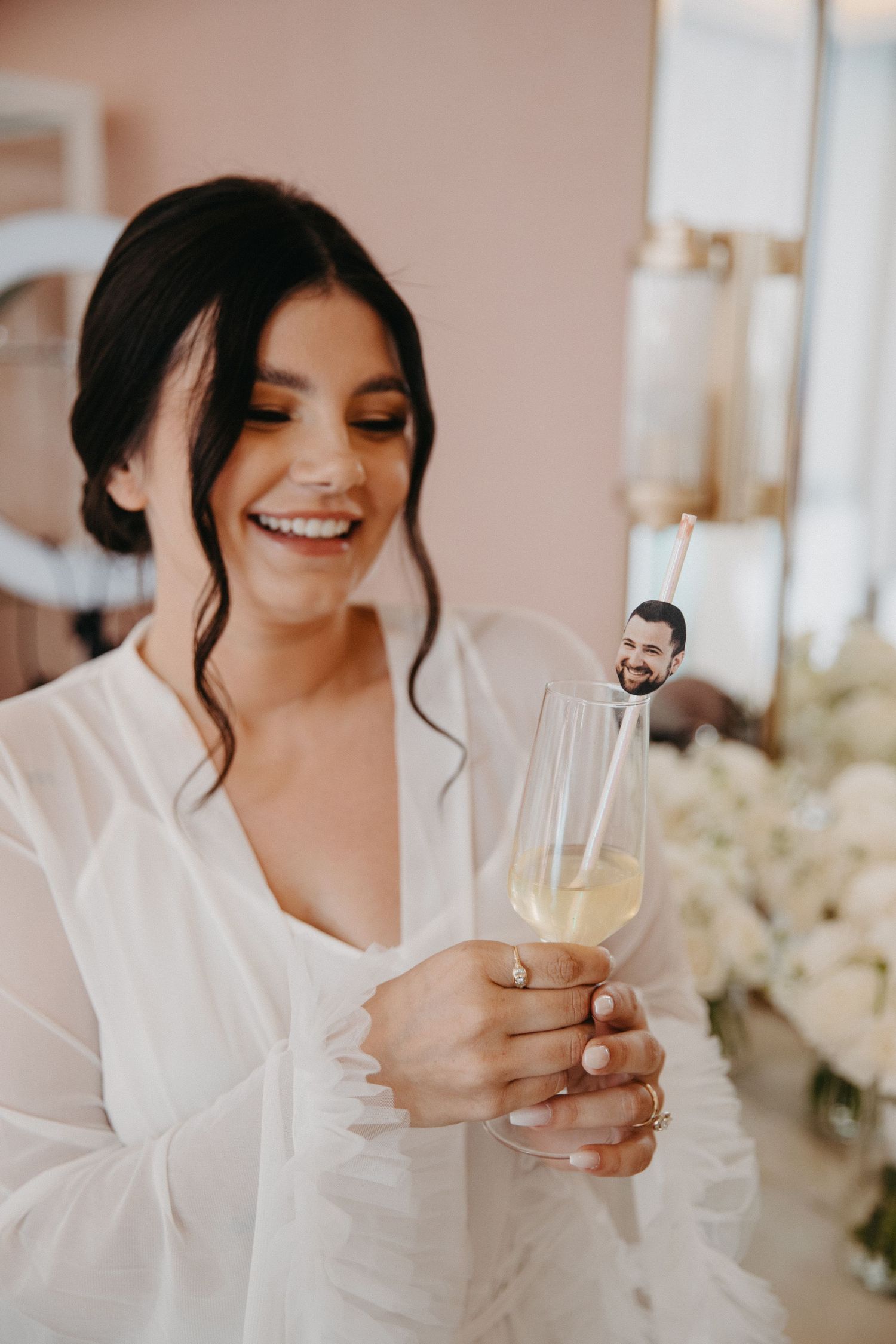 Bride in white robe holds champagne glass with decorative drink stirrer during wedding preparations.