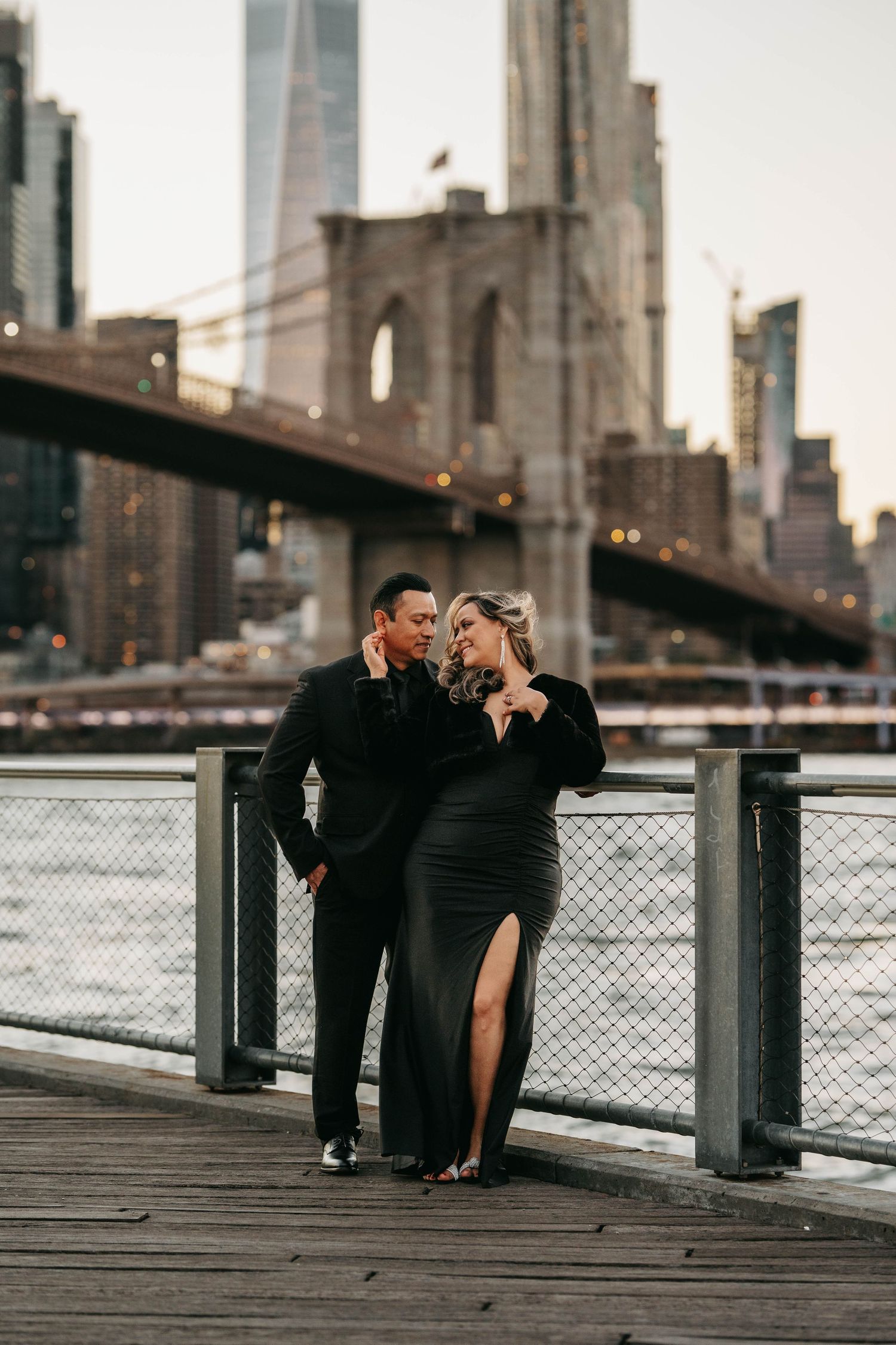 Romantic couple in formal black attire posing on Brooklyn Bridge waterfront with Manhattan skyline at dusk.