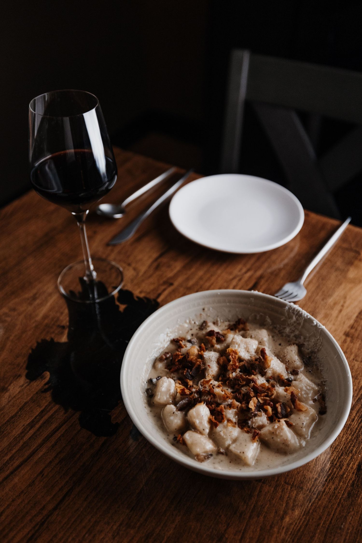 Bowl of gnocchi with dark sauce and glass of red wine on rustic wooden table.