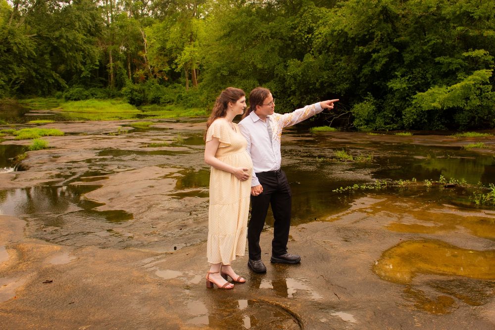 Pregnant couple standing in a shallow rocky stream, man pointing into lush green forest during outdoor maternity photoshoot.