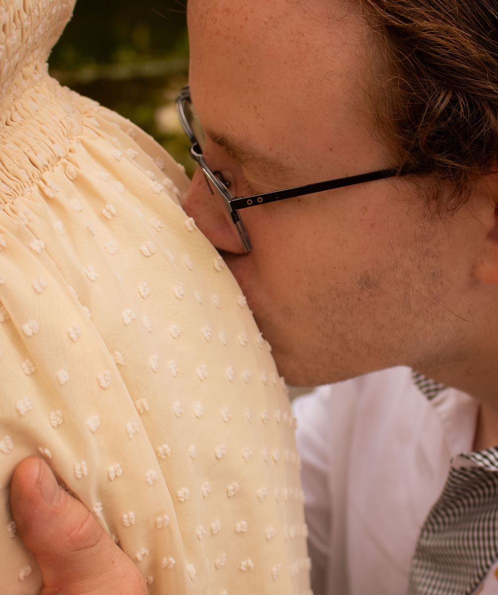 A man with glasses tenderly kisses a pregnant woman's belly while gently holding her cream dotted dress.