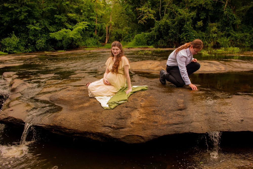 Pregnant woman in green dress sits on rock by waterfall as man crouches nearby in a lush forest setting.
