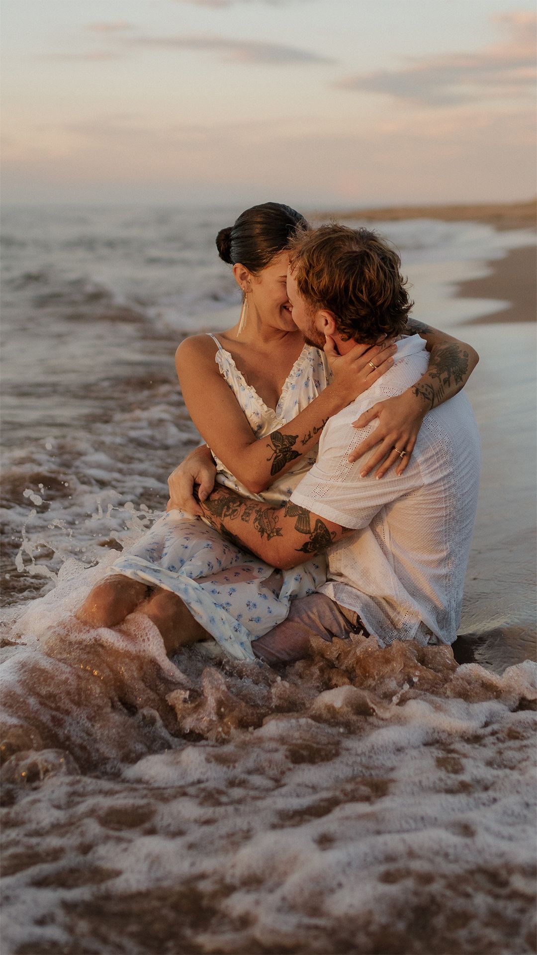 Couple embraces on sandy beach at sunset with waves washing around them.