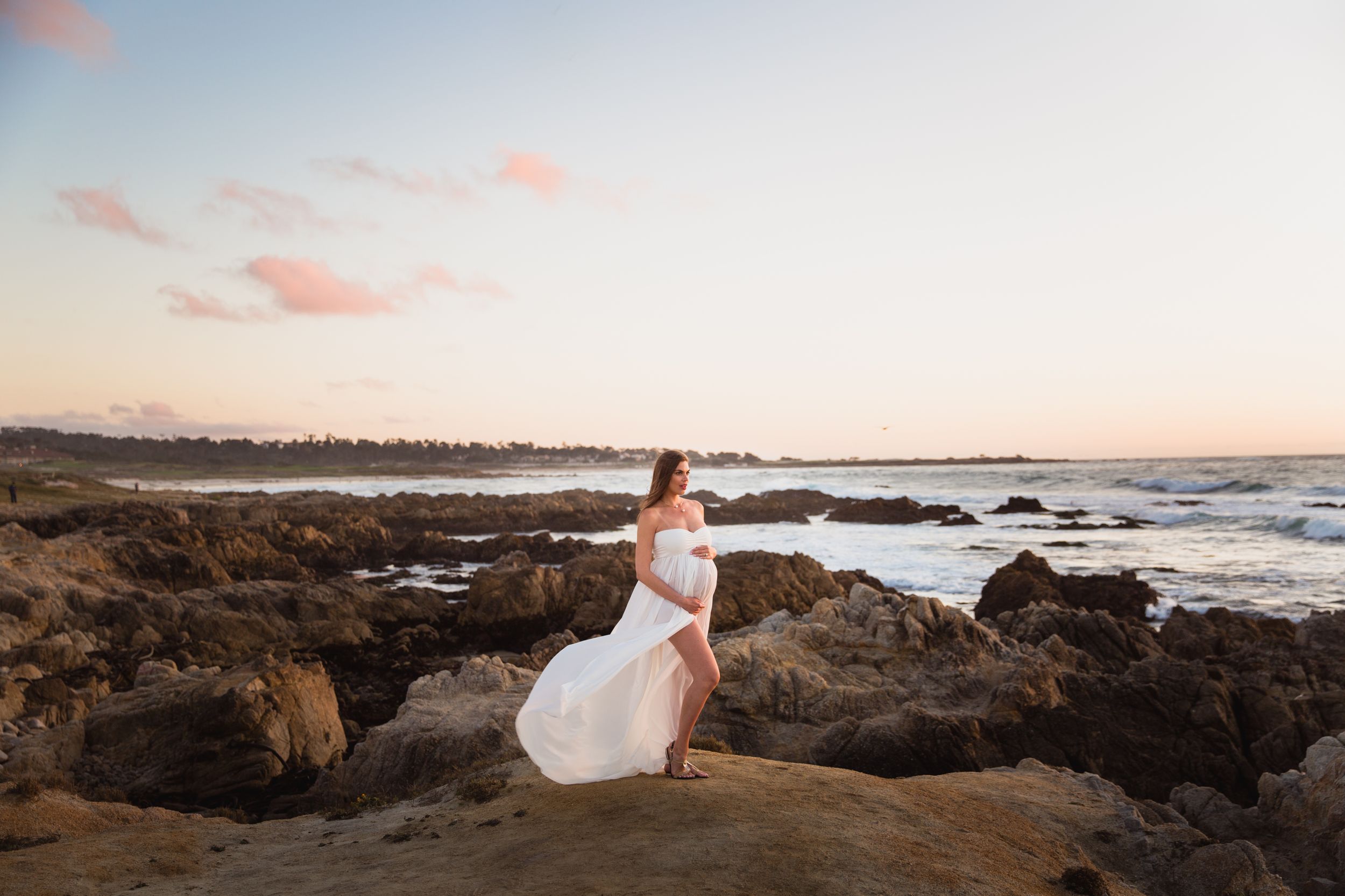 A pregnant woman in a flowing white dress stands on rocky coastal cliffs at sunset overlooking the ocean waves in Pacific Grove.