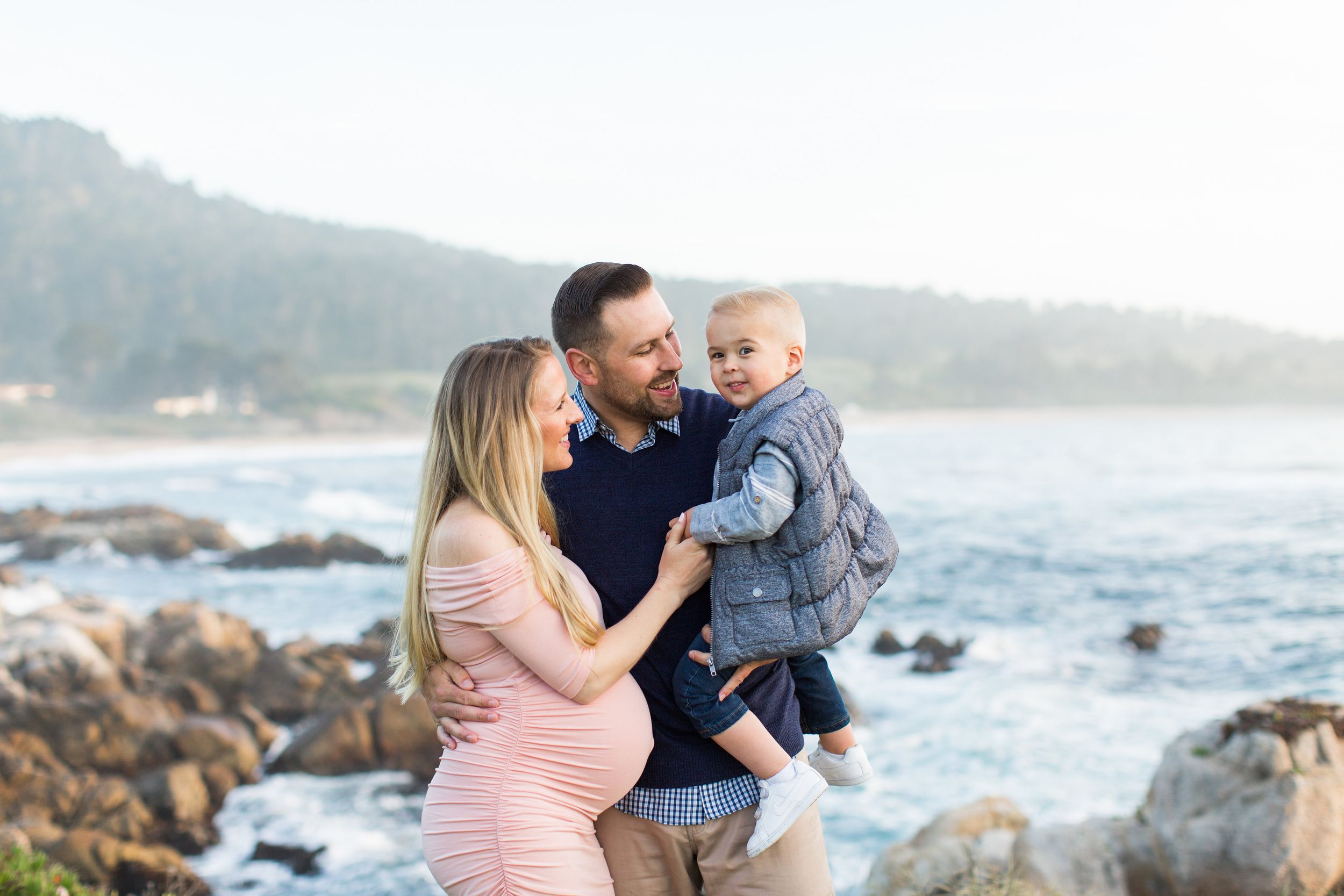 A family shares a tender moment together on a rocky beach at sunset with coastal mountains in the background in Carmel, CA with Rachel Zee.