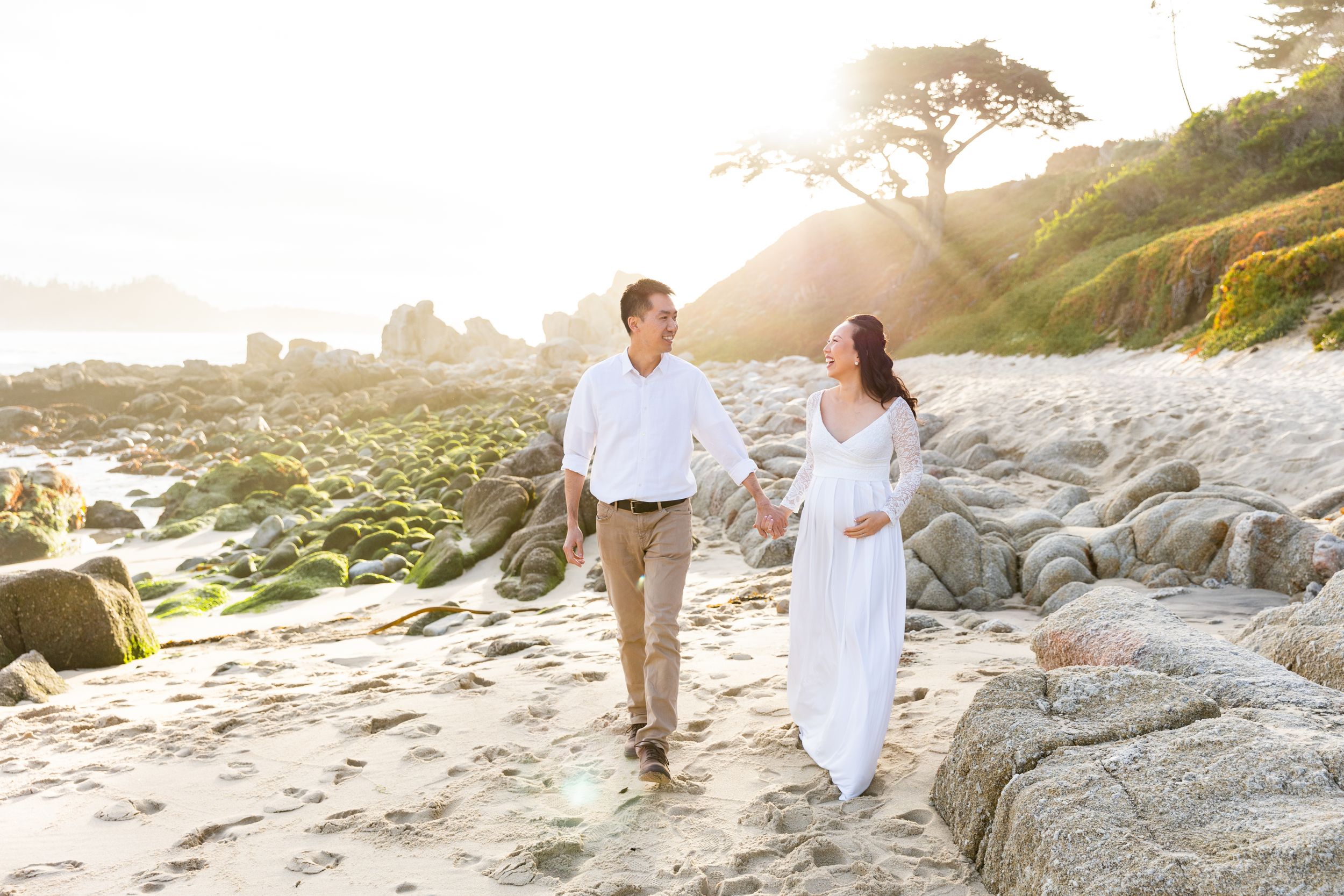 A couple walks hand in hand on a rocky beach at sunset with golden light filtering through coastal trees in Carmel, CA.