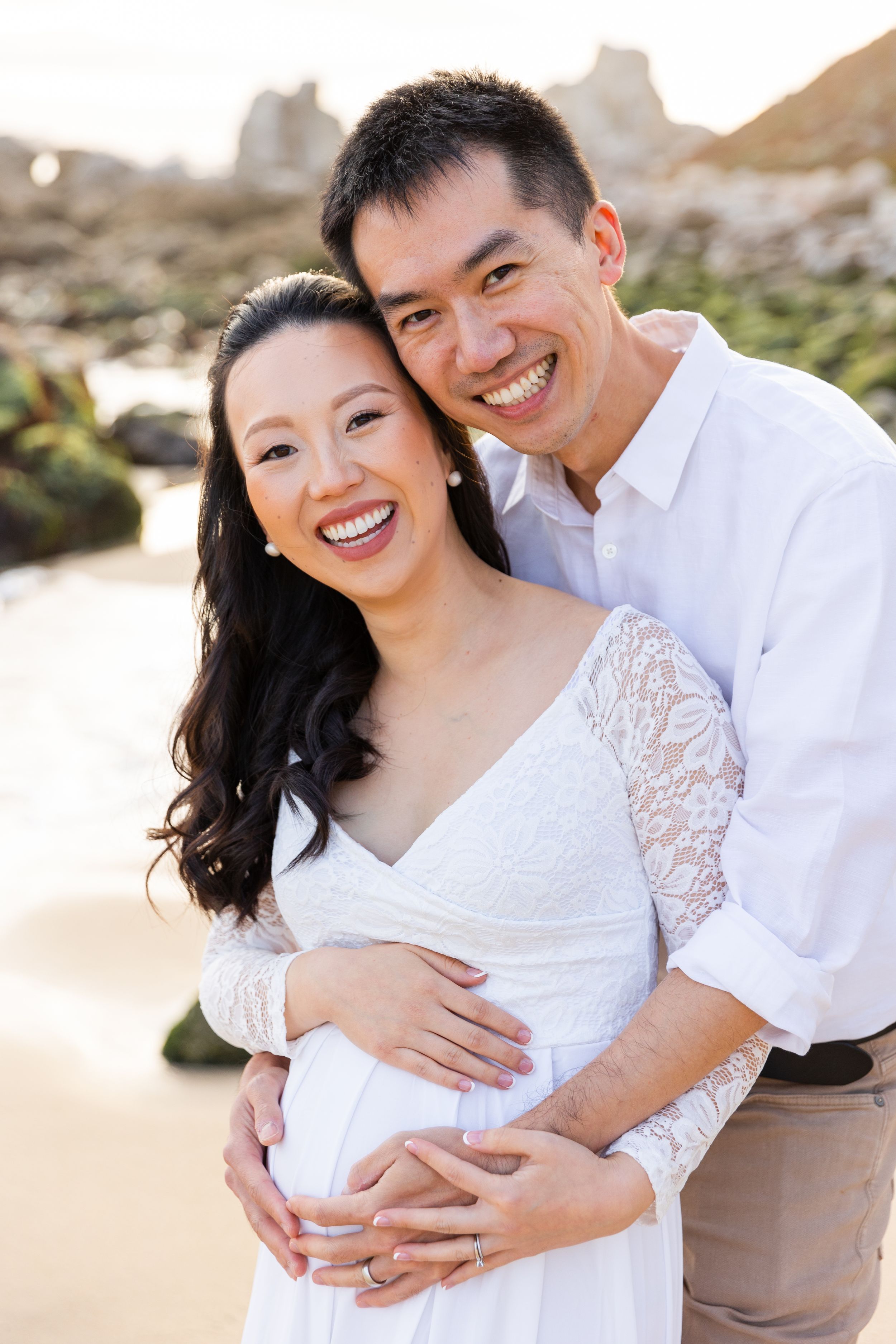 A couple in white clothing embrace and smile warmly during a maternity photoshoot on a beach at sunset during babymoon in Carmel.