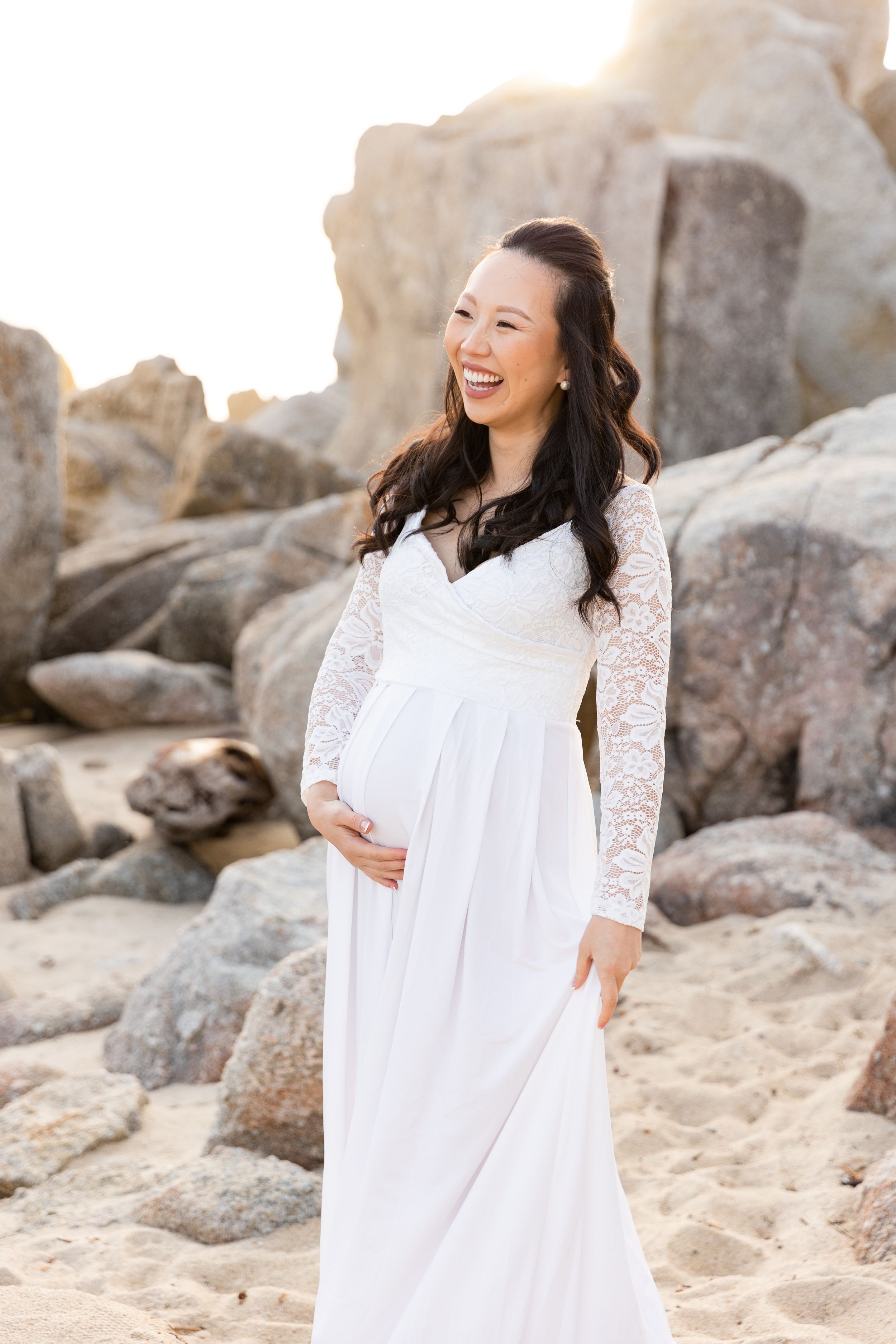 Beautiful maternity photo session features an expectant mother in a white lace dress laughing on a rocky beach by Rachel Zee Photography.