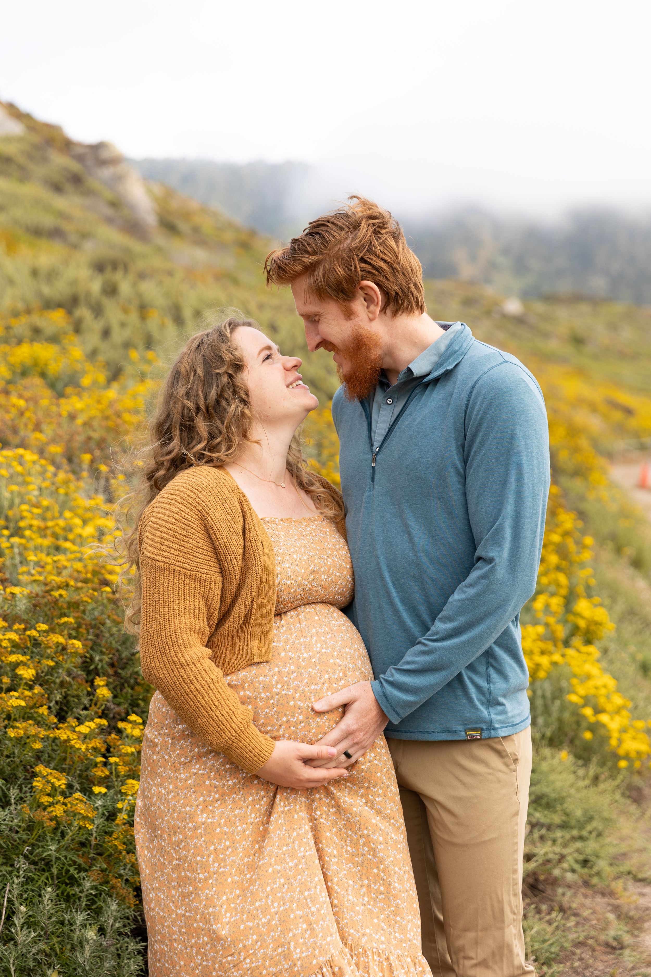 A couple stands together in a field of yellow wildflowers, for their maternity session during their babymoon in Carmel, California.