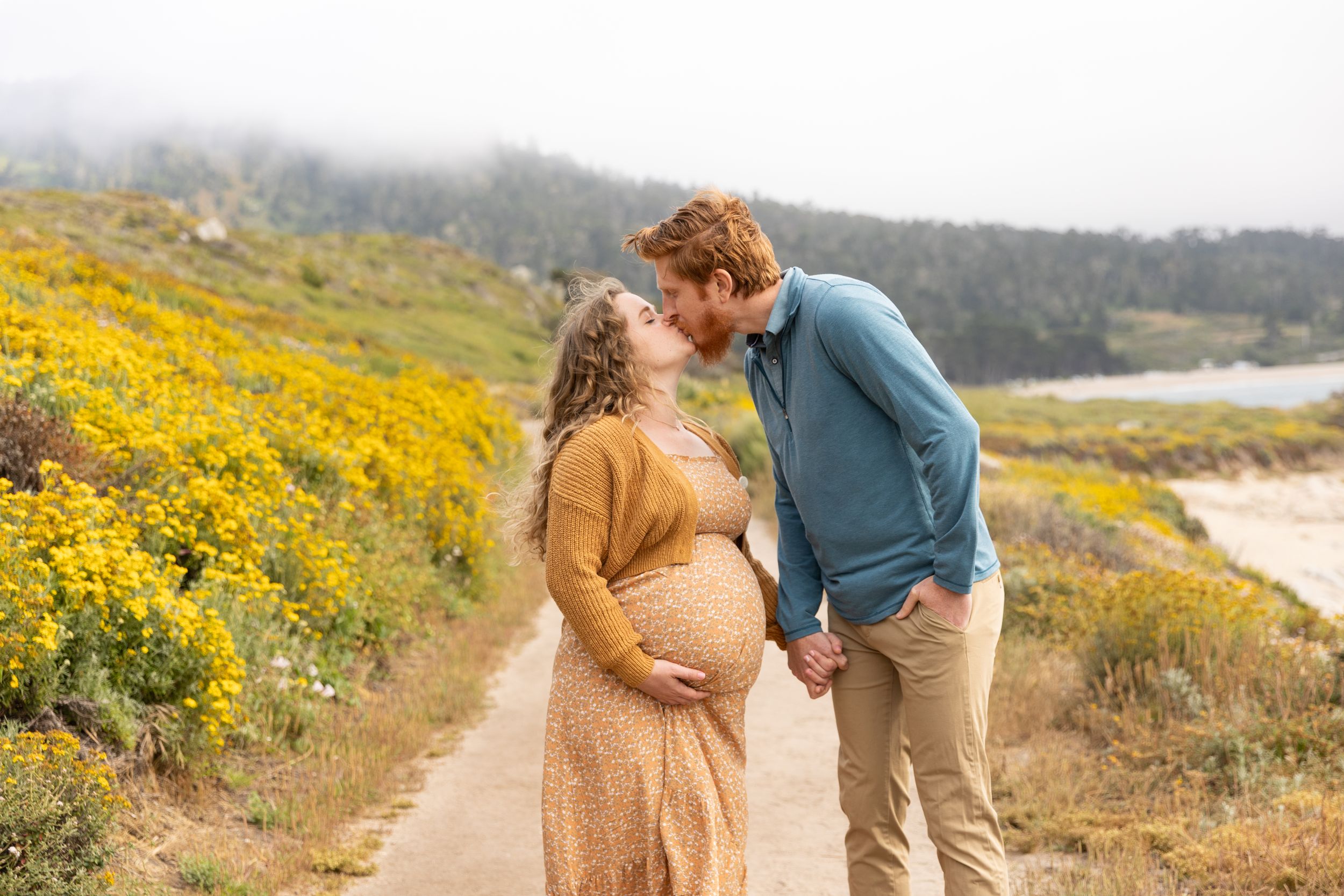 A couple embraces on a trail surrounded by vibrant yellow wildflowers along a misty coastal hillside on their babymoon in Carmel.