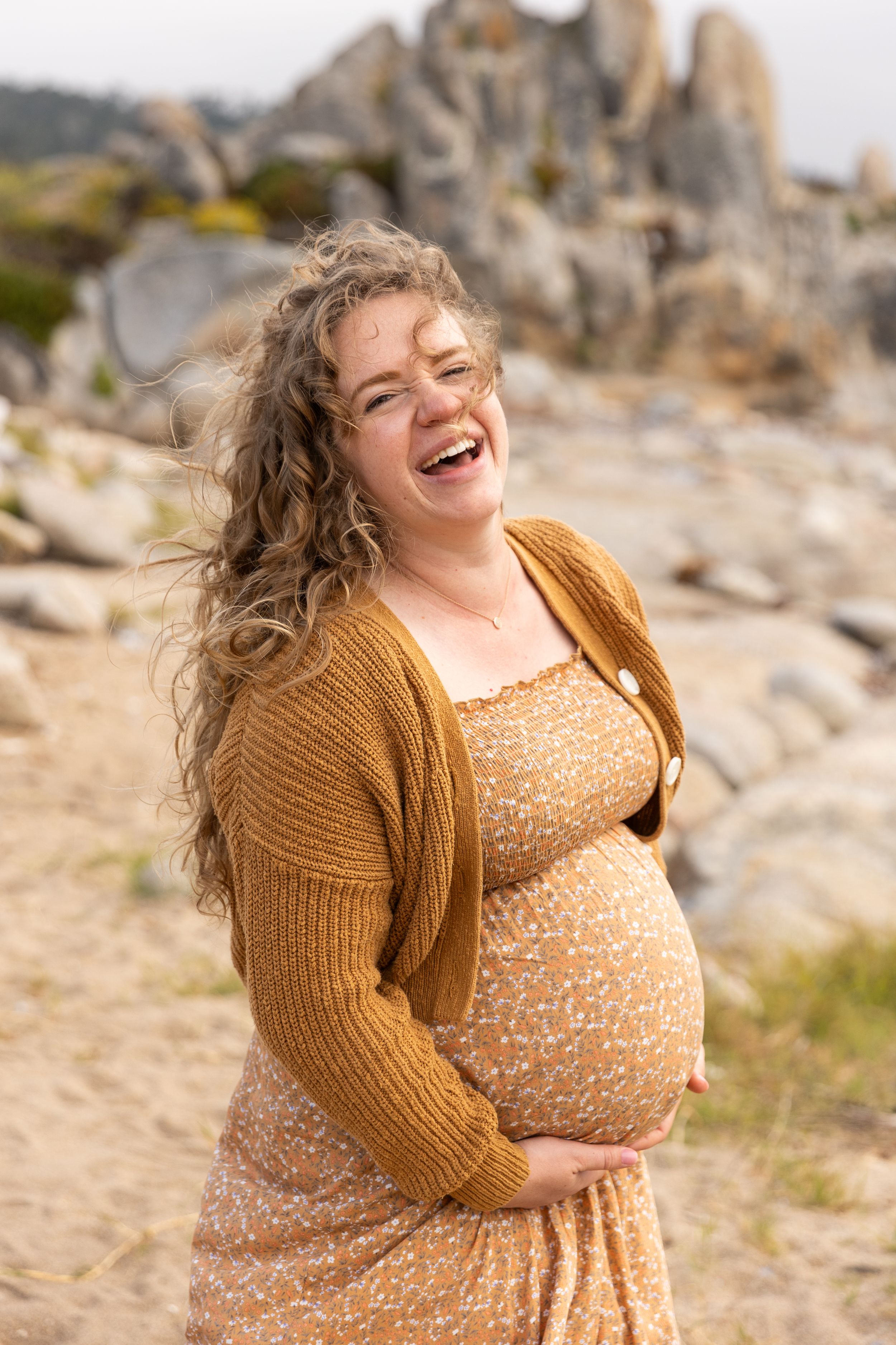 A joyful expectant mother in a floral dress and mustard cardigan laughs while cradling her belly against rocky scenery in Carmel, CA.