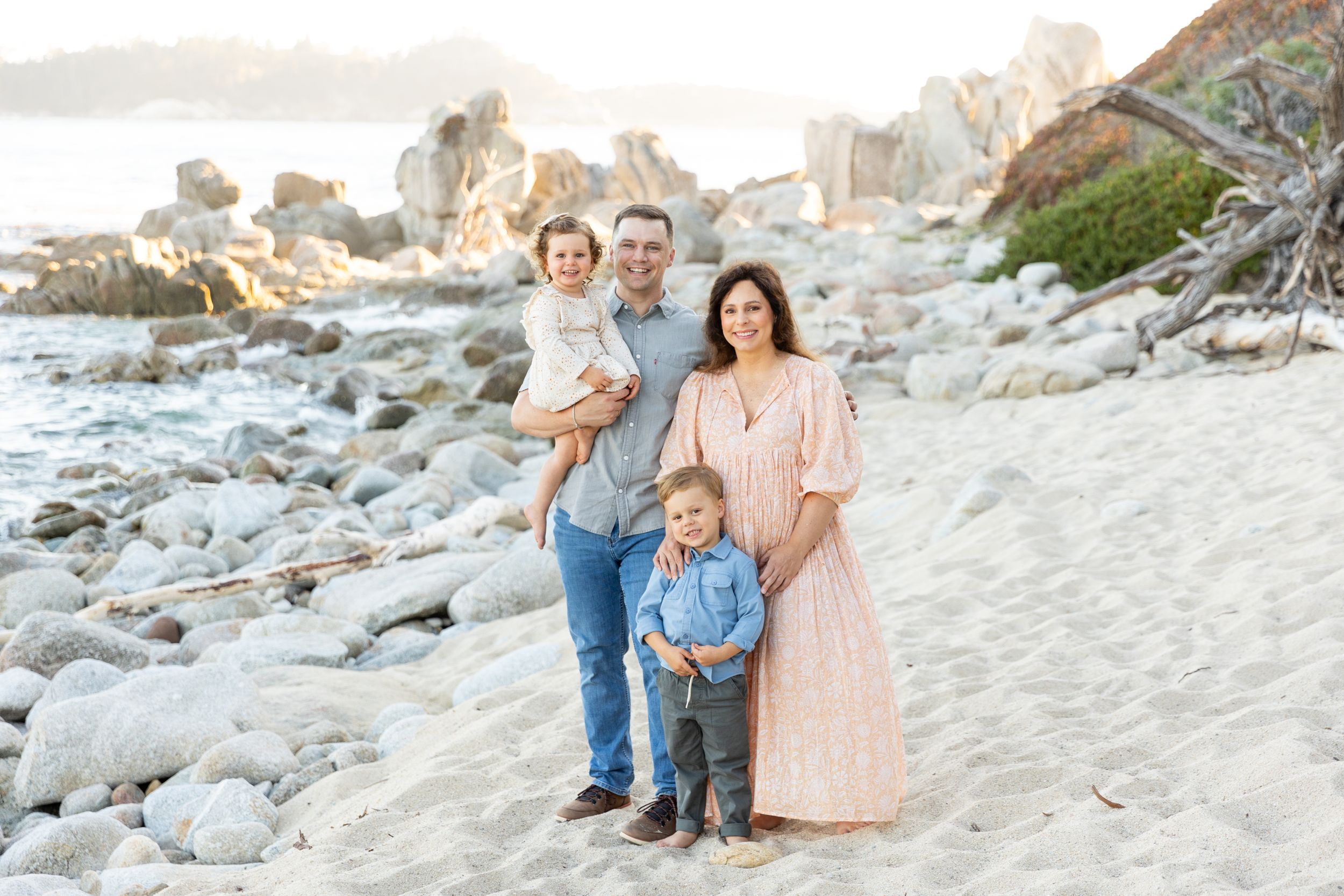 A family stands together on a rocky beach at sunset with ocean cliffs visible in the background in Carmel, CA.
