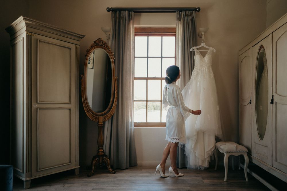 A white wedding dress hangs by a window in an elegant dressing room with antique furniture.