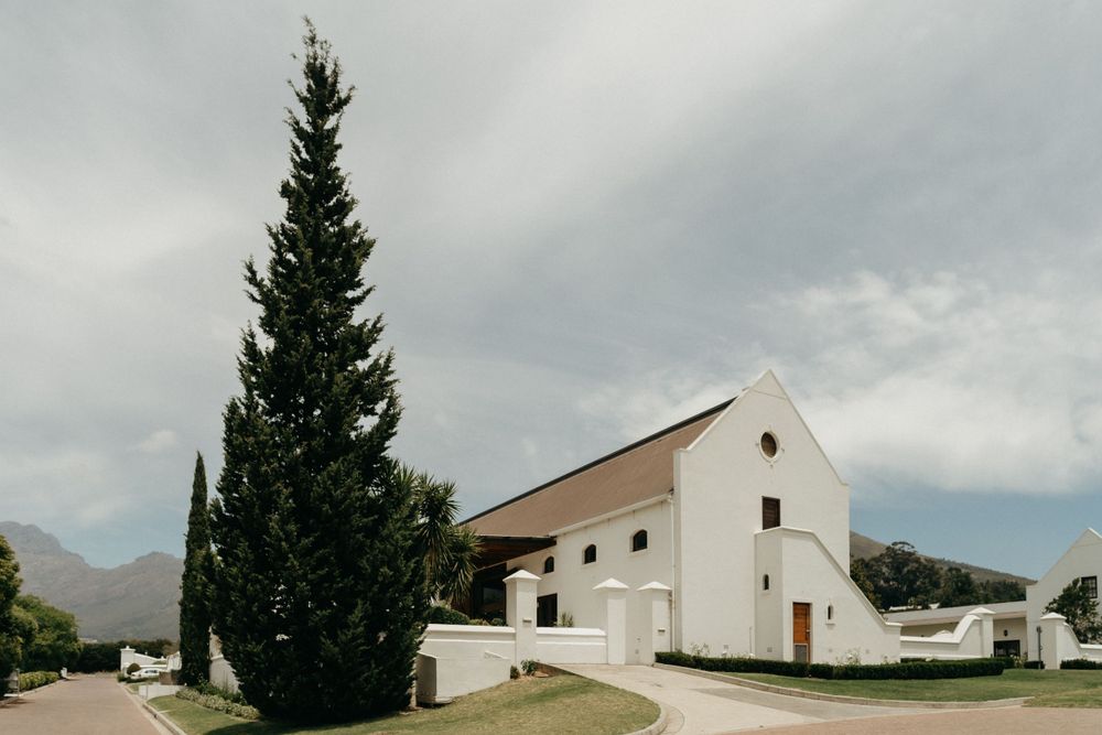 White church building with cypress tree under cloudy skies in mountain setting.