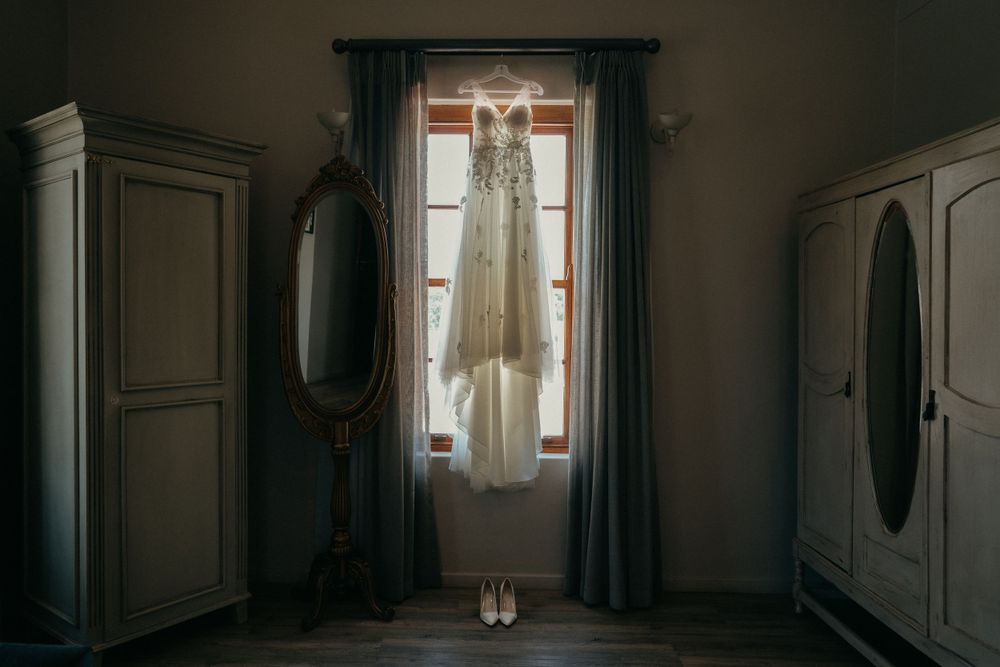 An elegant white wedding dress hangs in front of a window with dark curtains in a rustic room.