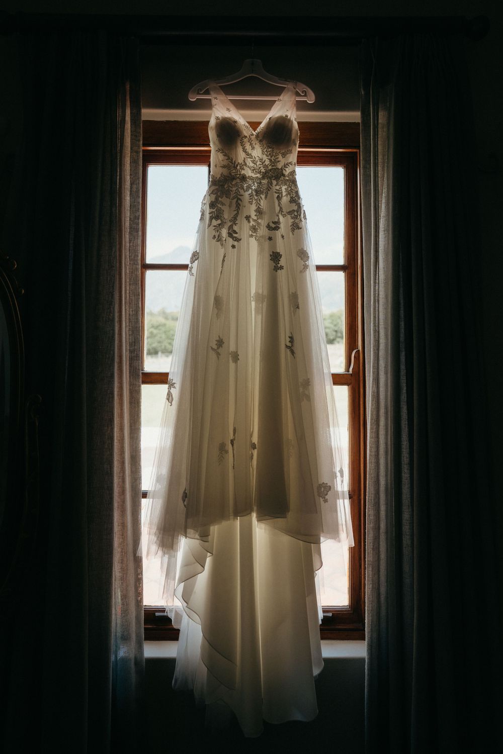 A delicate lace wedding gown with floral details hangs backlit against a window between dark curtains.