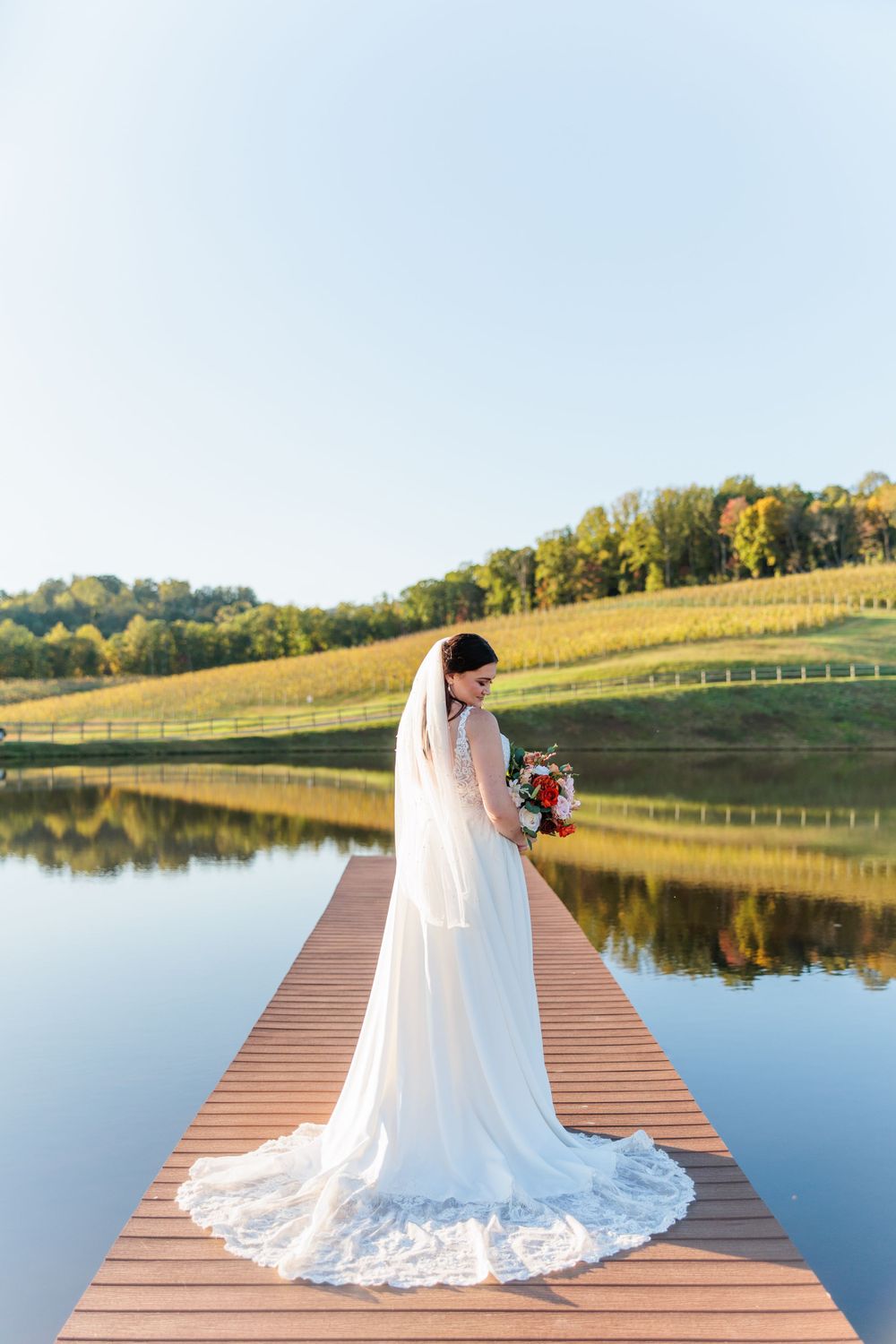 Bride in lace gown stands on wooden dock overlooking serene lake and autumn landscape.