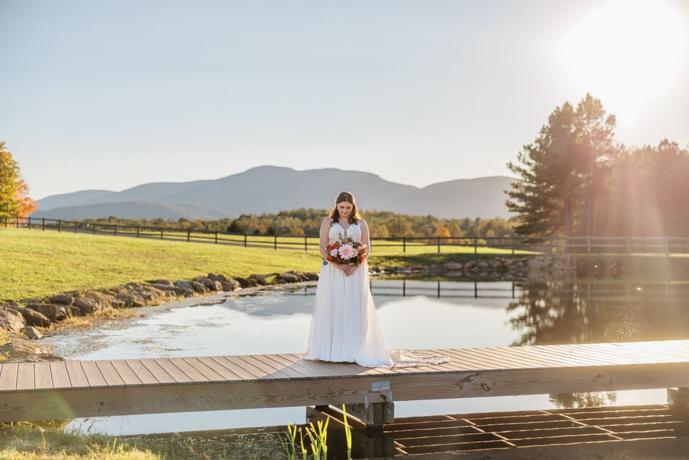 Wedding photo on wooden dock overlooking tranquil pond with mountains and sunset in background.