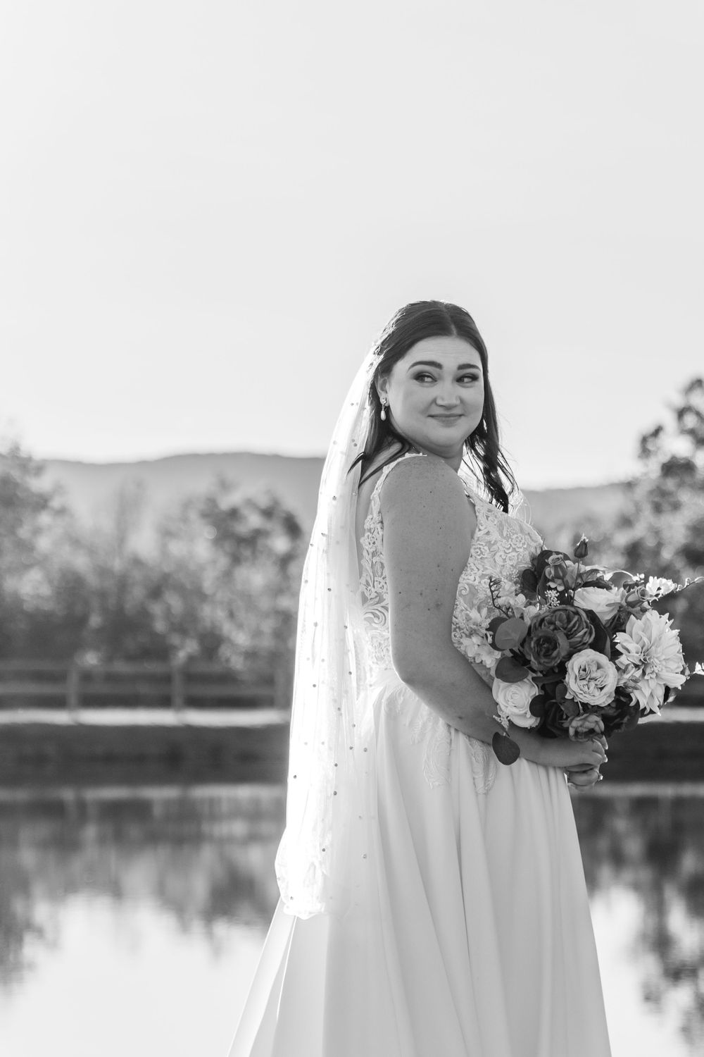 Black and white bridal portrait by lakeside with mountain scenery and floral bouquet.