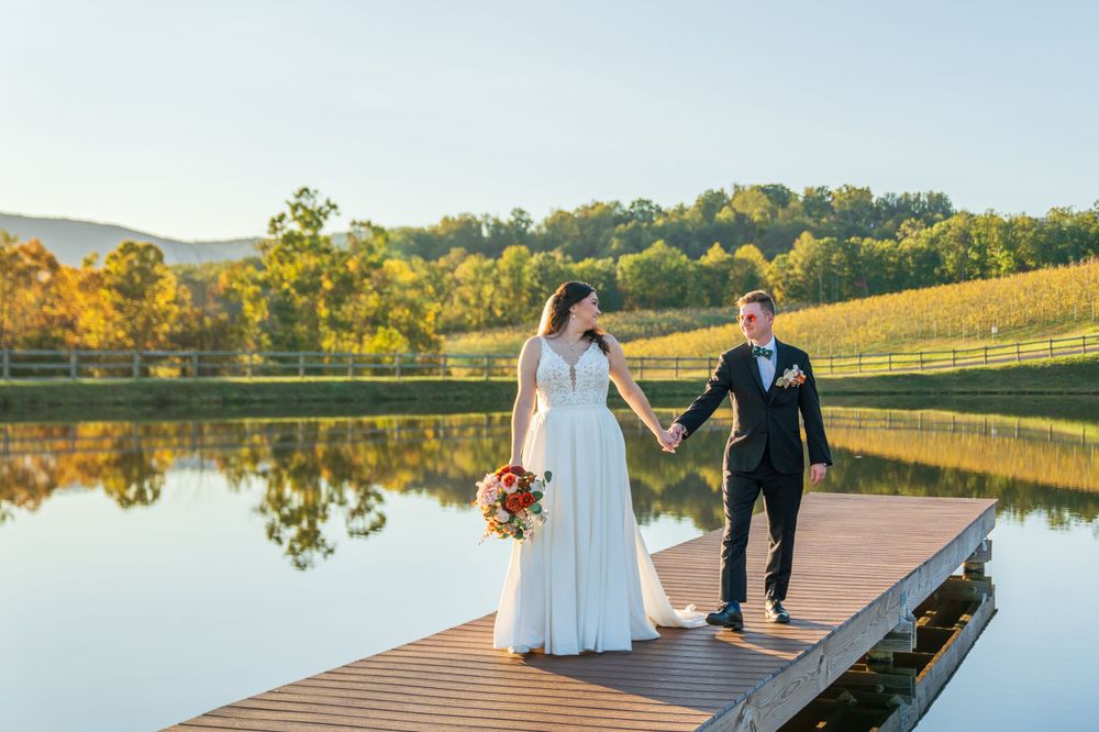 Wedding couple walking on wooden dock over reflective pond with autumn mountain backdrop.
