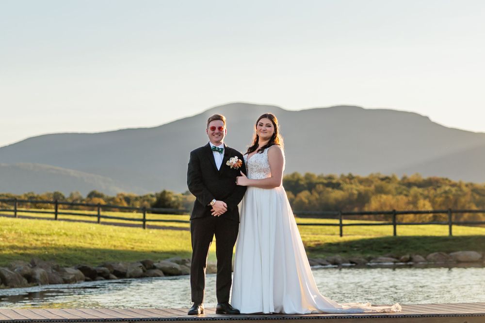 Newlyweds pose together on dock with mountain vista and grassy fields behind them.