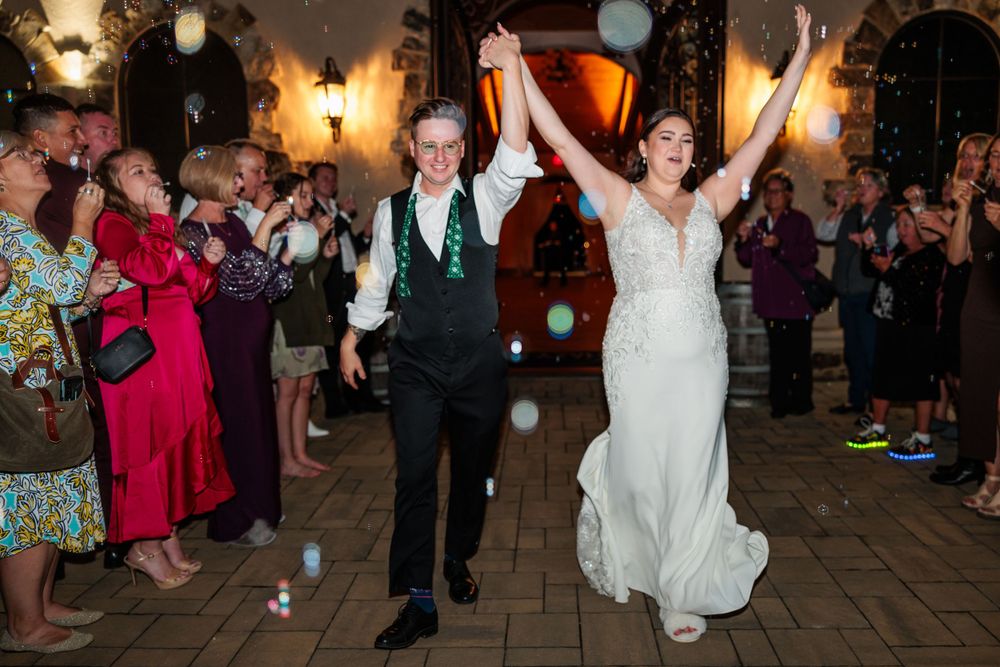 Newlyweds make their grand exit through a crowd of cheering guests with arms raised in celebration at a dimly lit venue.