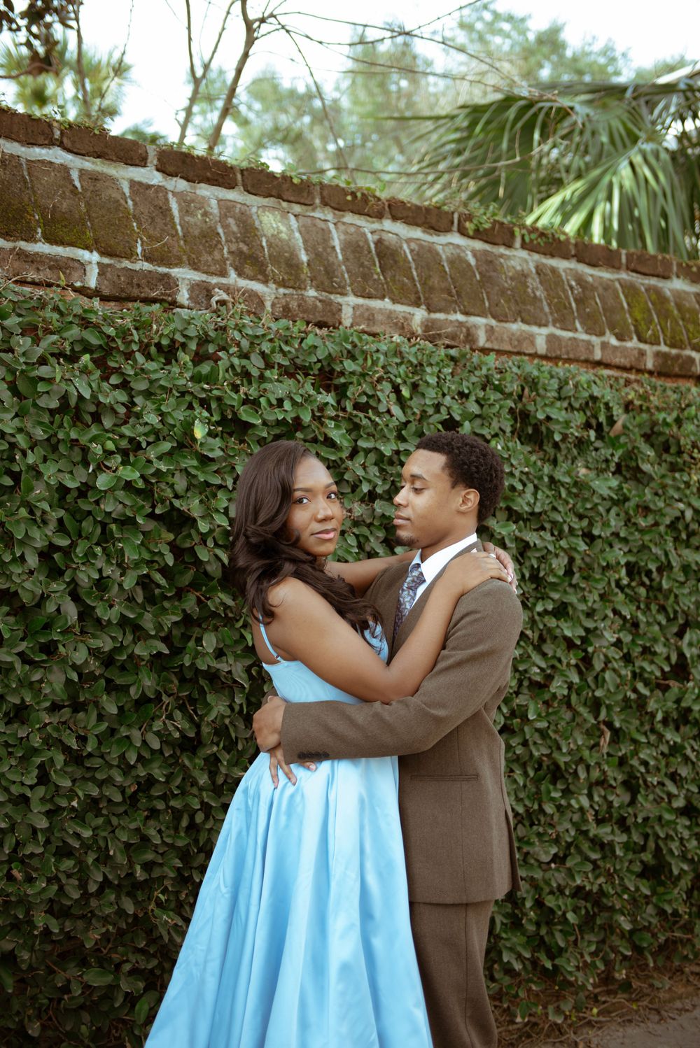 A couple embracing, the girl is looking at the camera while the guy is looking at her. There is a brick wall behind them with greenery.