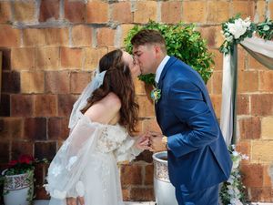 A bride and groom share their first kiss as newlyweds against a rustic brick wall decorated with greenery and white flowers.