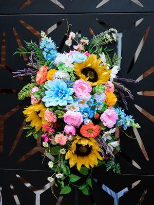 Colorful floral arrangement with sunflowers, pink roses, and blue hydrangeas against a dark background with decorative accents.
