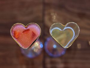 Two heart-shaped glass containers with pink and clear liquids photographed against a dark wooden background.