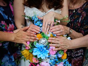 Multiple hands with colorful manicures rest on a vibrant bouquet of blue, yellow and pink flowers.