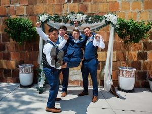 A group in navy suits poses playfully under a floral wedding arch against a rustic brick wall.