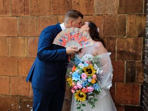 A couple shares a romantic kiss against a rustic brick wall while holding a vibrant multicolored wedding bouquet.